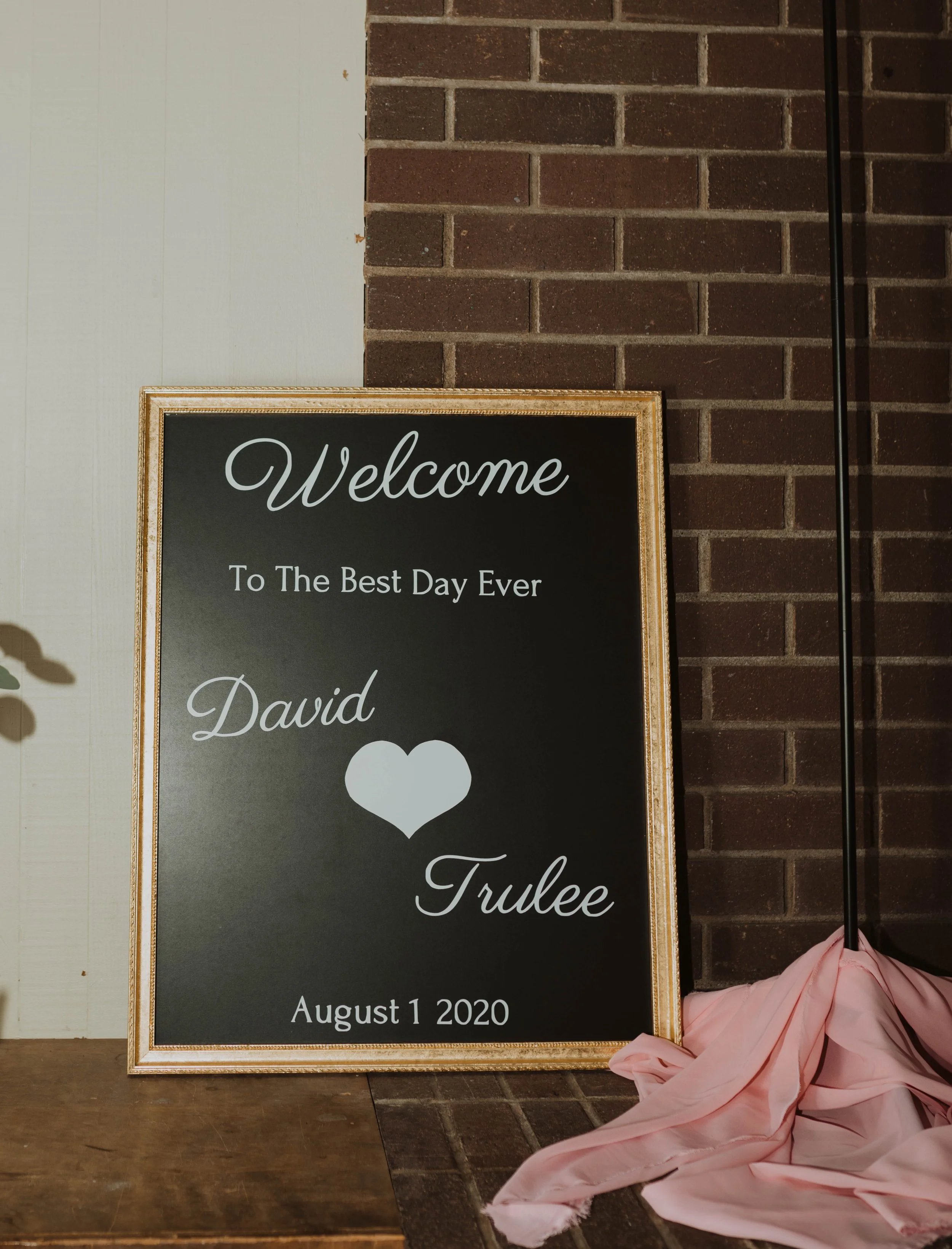 Welcome signboard for a wedding with the names David and Trulee, date August 1, 2020, against a brick wall and decorated with pink fabric. Seattle, WA wedding photography.