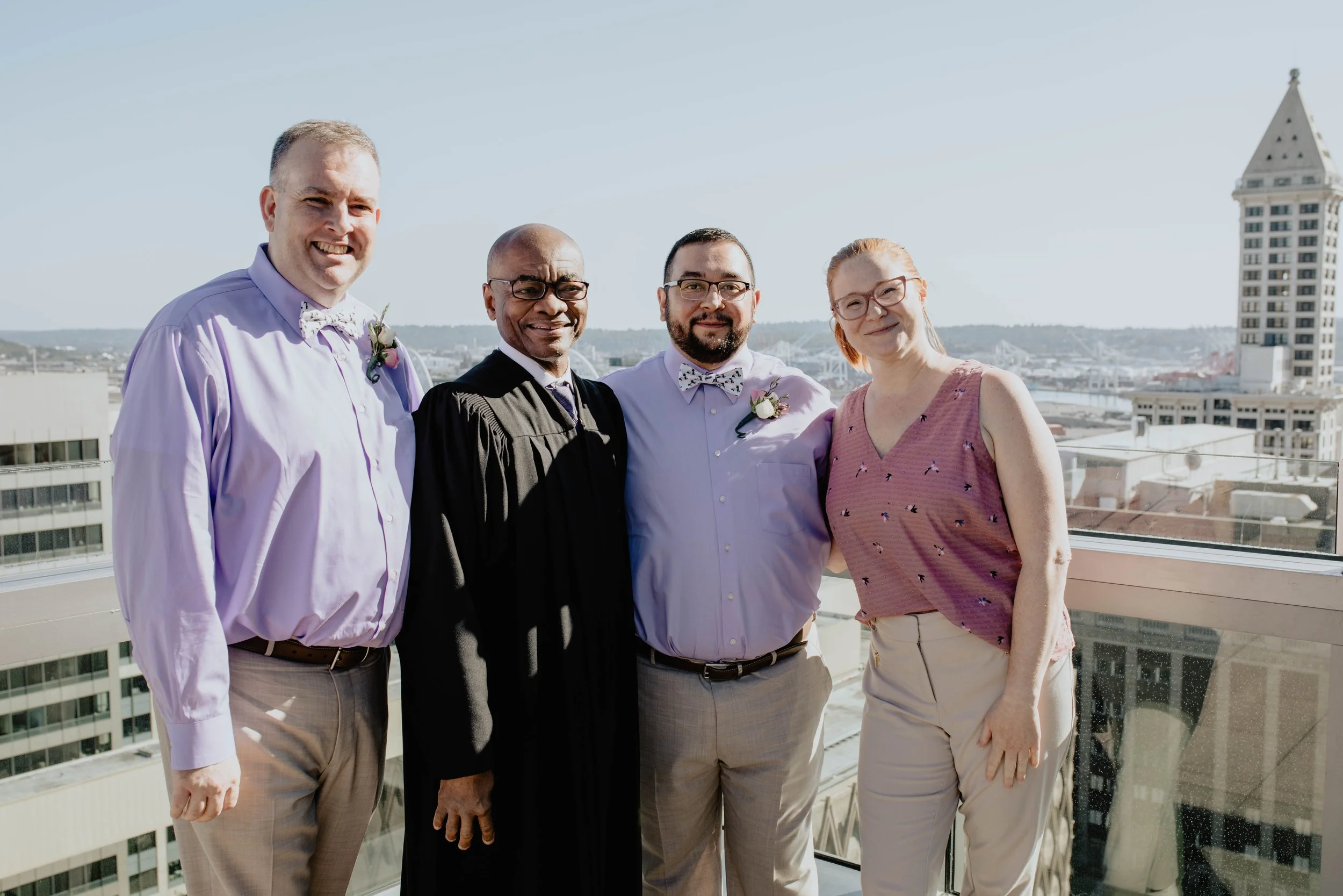 Group of five people standing on a balcony overlooking a cityscape with a tall building in the background, dressed in formal and semi-formal attire, smiling at the camera. Seattle Municipal Courthouse wedding photography.
