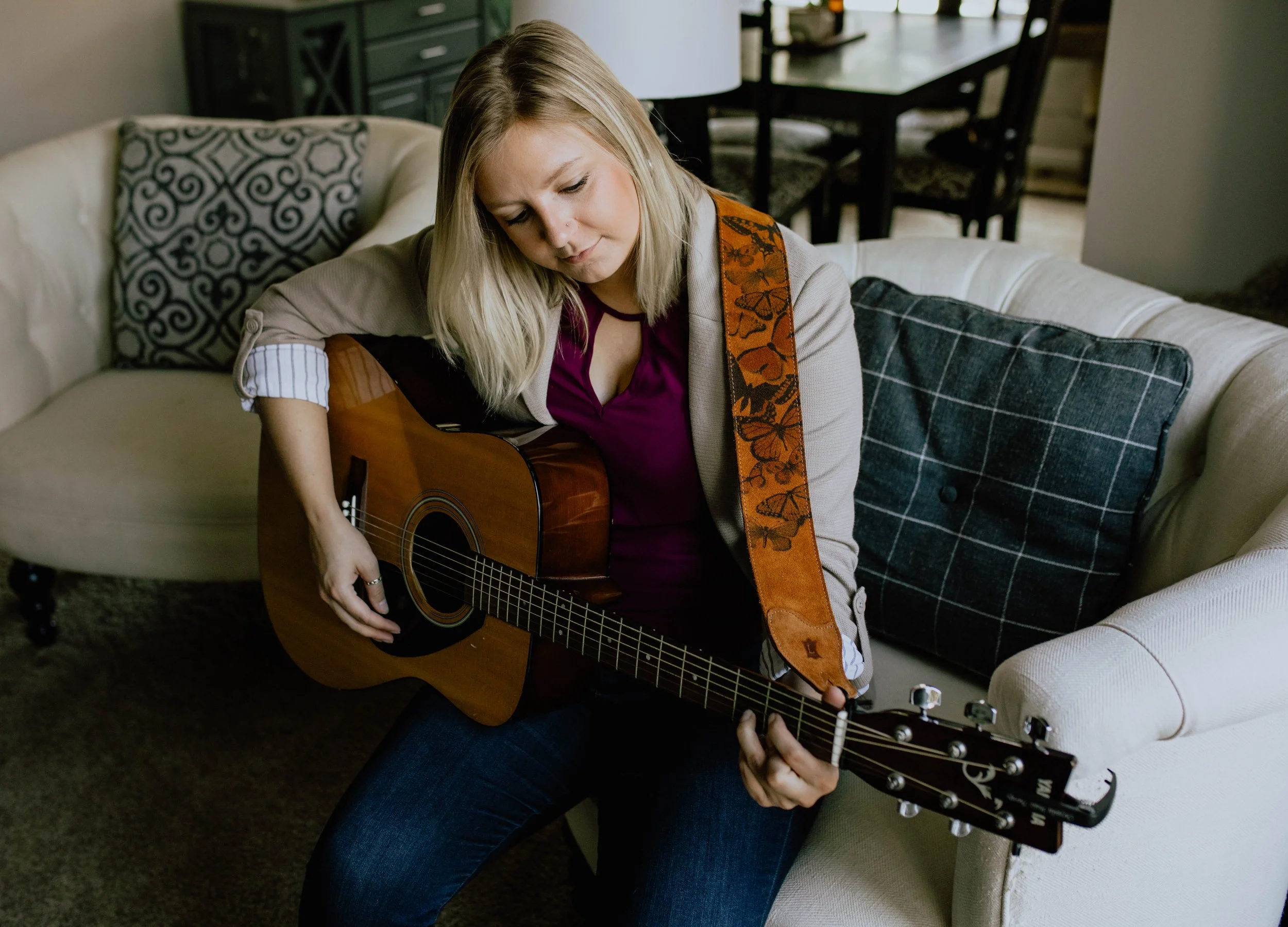A woman with blonde hair sitting on a cream-colored couch, playing an acoustic guitar. She is wearing a beige blazer with patterned elbow patches over a purple top, with her left hand on the fretboard and her right hand strumming.