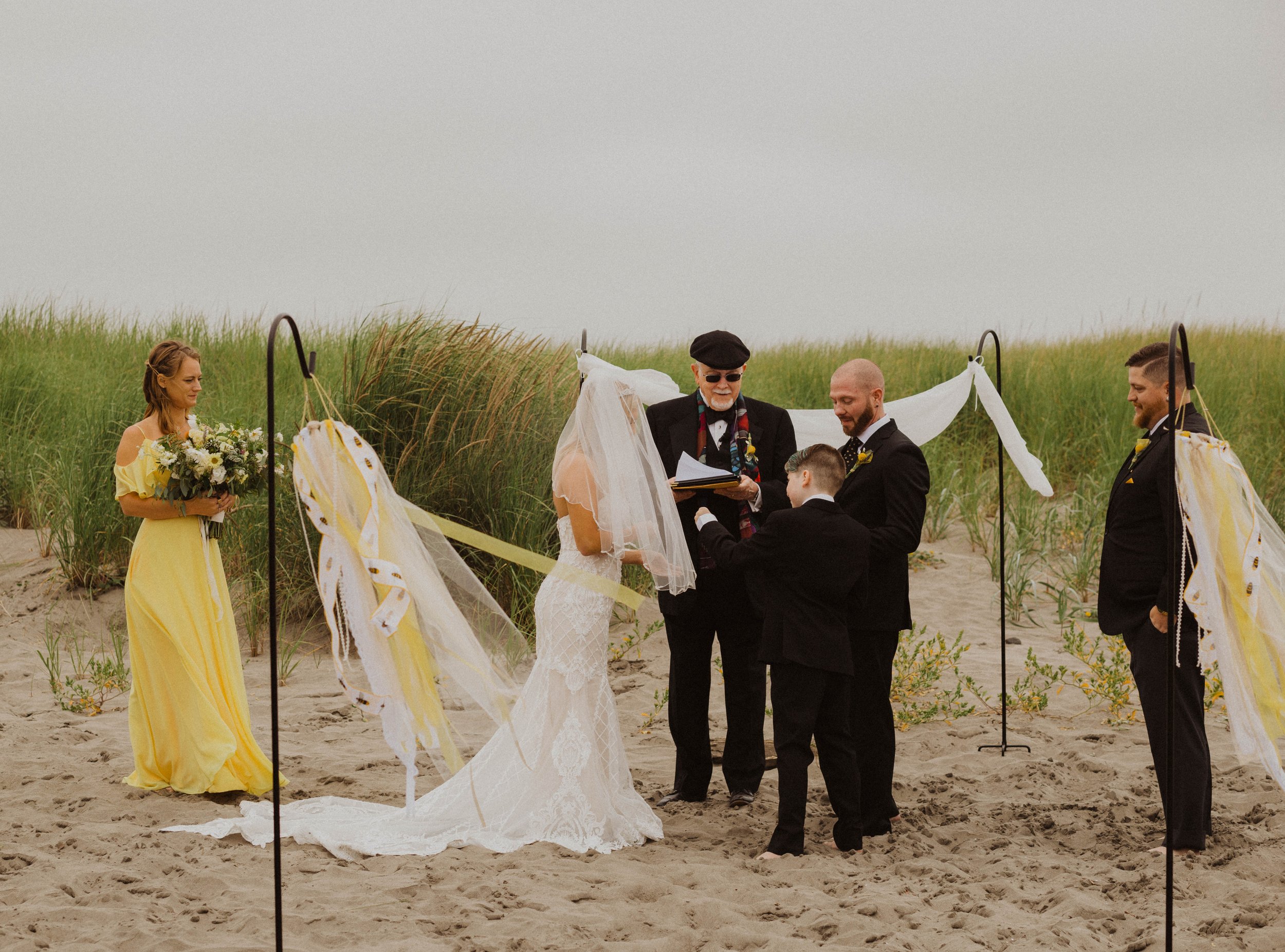 A beach wedding ceremony with a bride and groom, officiant, two groomsmen, and a bridesmaid, surrounded by grassy dunes. Long Beach, WA wedding photography.