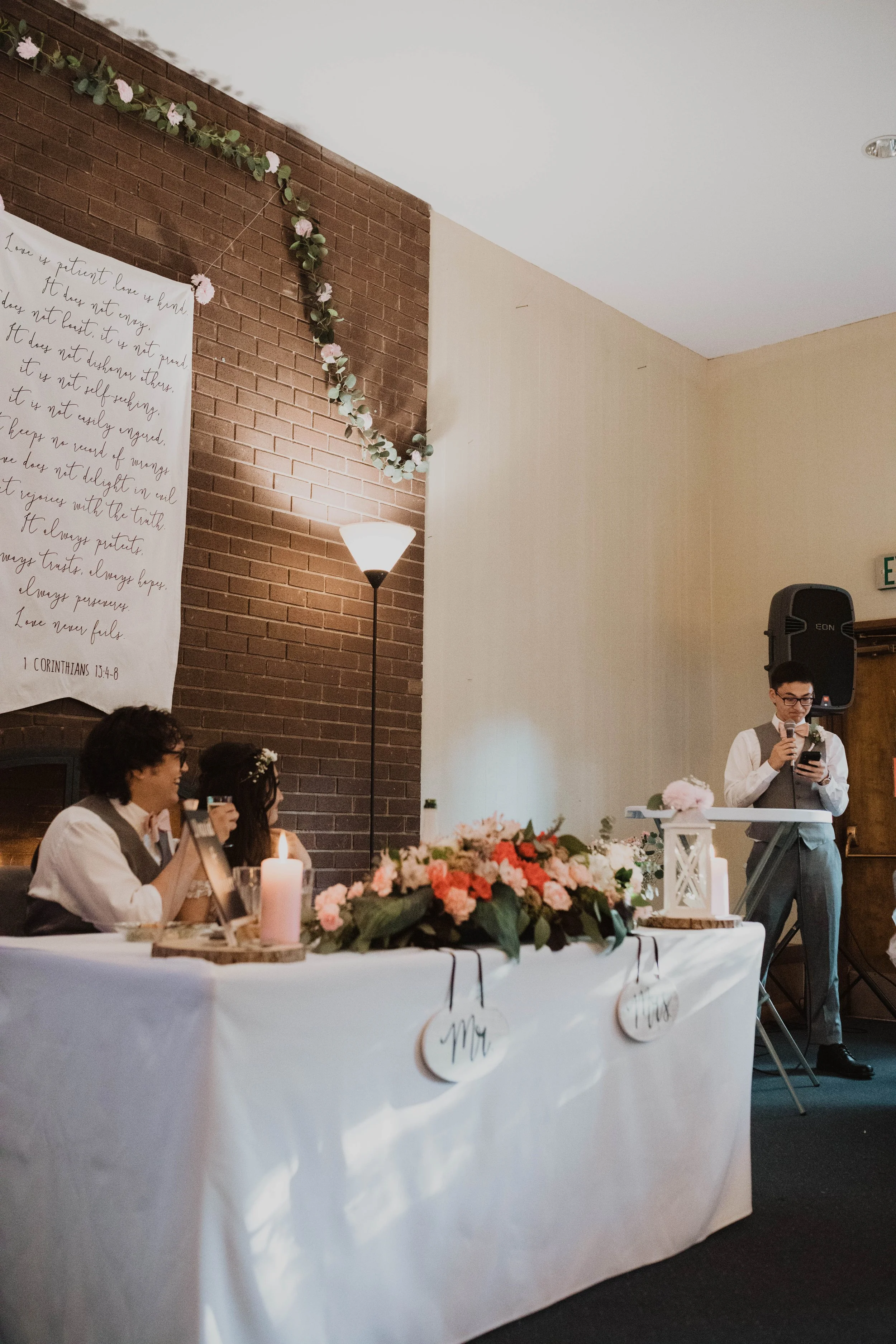 A wedding reception with a table decorated with flowers and candles. Two people are seated at the table, the groom and bride, both dressed in wedding attire. A man is standing at a lectern giving a speech, holding a microphone and a phone. The backgr