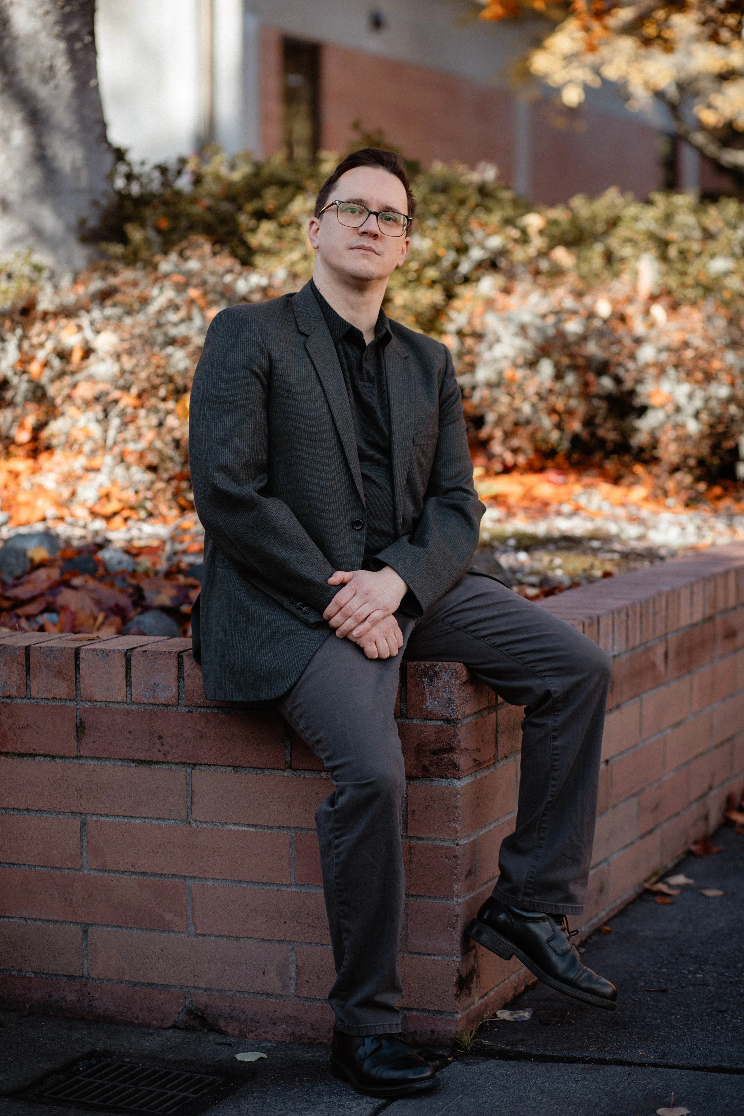 A man in a dark gray suit and black shirt sits on a brick ledge outdoors during autumn, with colorful fallen leaves and bushes in the background. Seattle professional head shot photography
