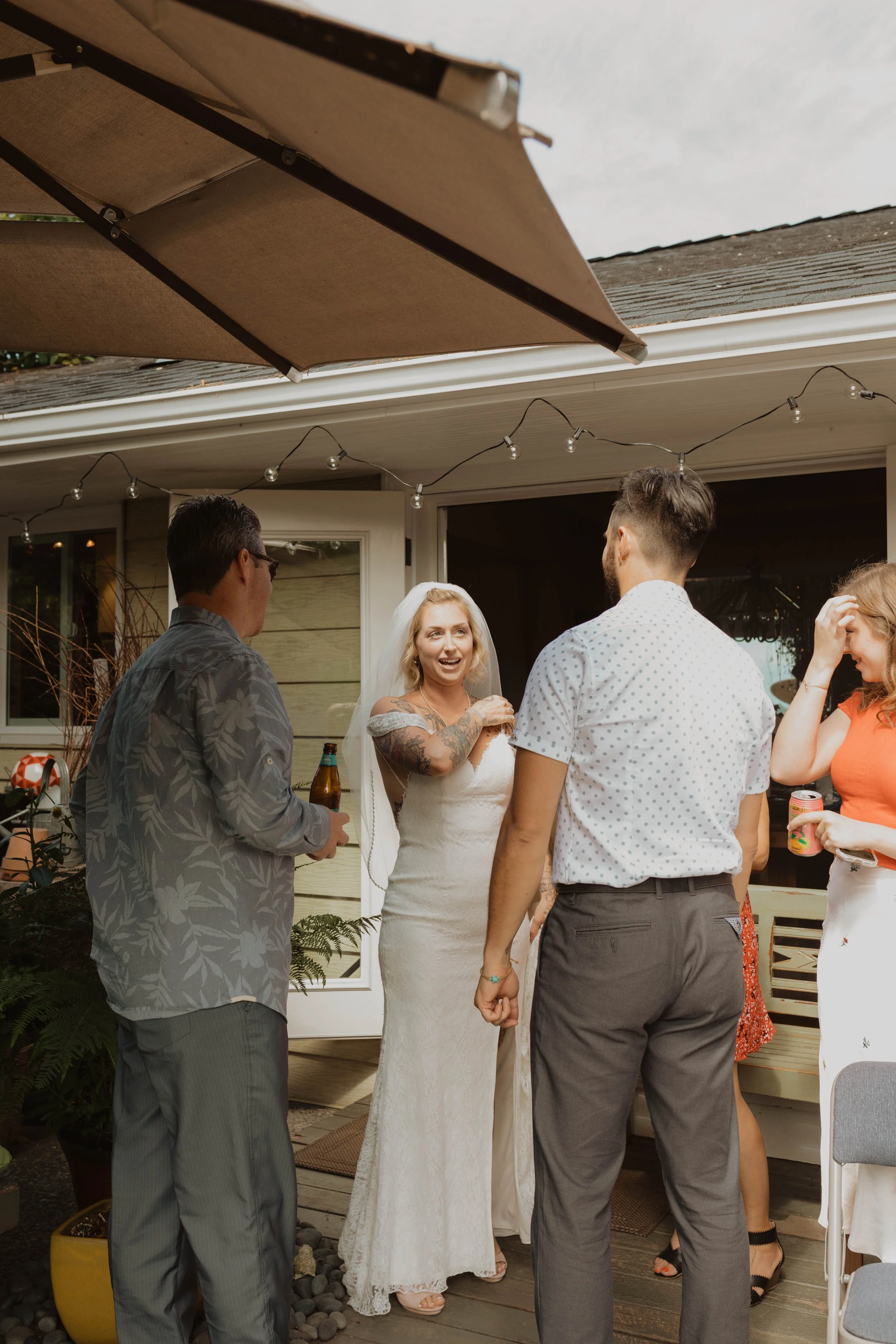 A group of people at a wedding reception outside, including a woman in a white dress with tattoos, talking and smiling with others near a house with string lights and an umbrella overhead. Seattle, WA wedding photography.