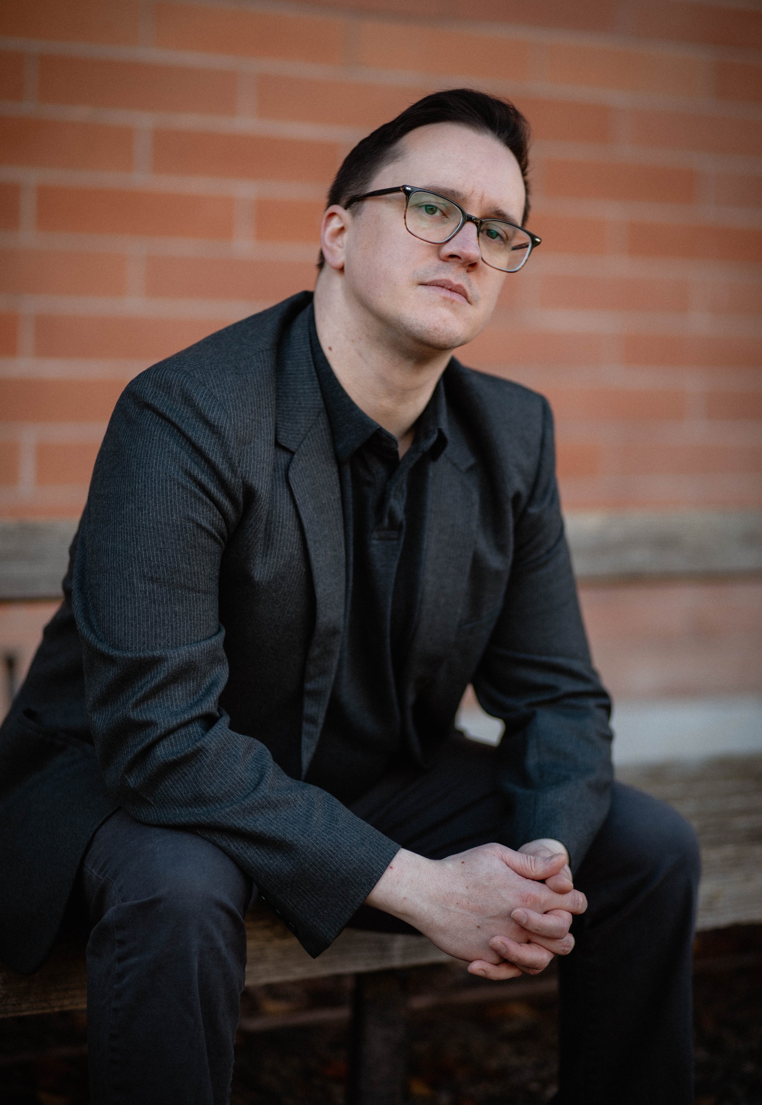 A young man with short dark hair and glasses, wearing a black blazer and shirt, sitting on a wooden bench outdoors against a brick wall, with a serious expression and hands clasped. Seattle professional head shot photography