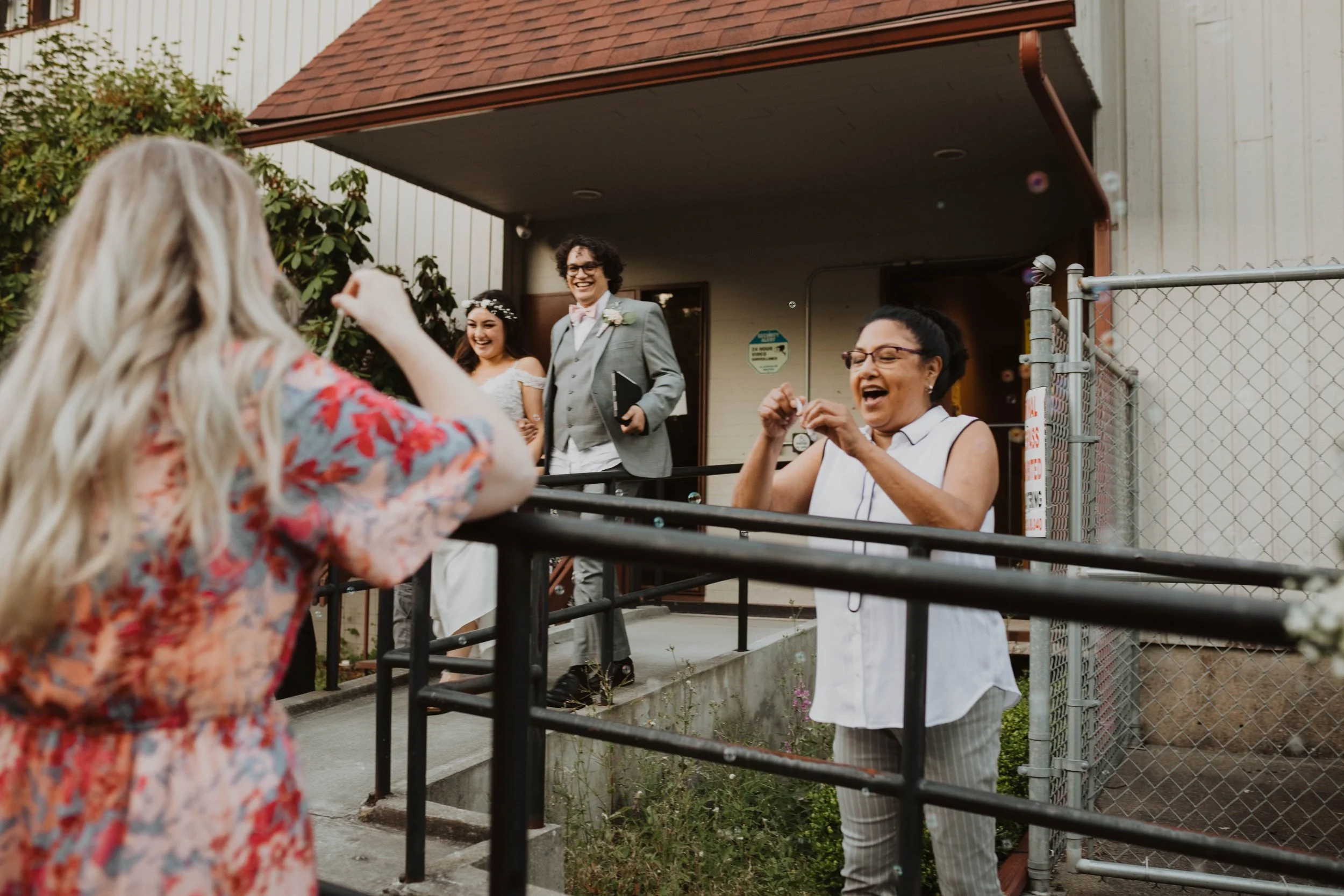 A wedding celebration with a bride, groom, and two women outside a building. The bride wears a white wedding dress and headpiece, the groom in a gray suit with a pink bow tie, and the two women are laughing and capturing the moment.