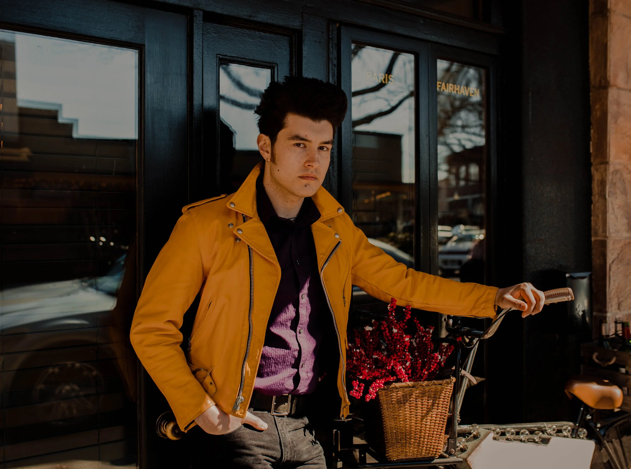 A young man with dark hair and a serious expression wearing a yellow jacket, standing outside a building with black doors and reflective windows, holding a bicycle with a basket of red berries. Seattle professional head shot photography