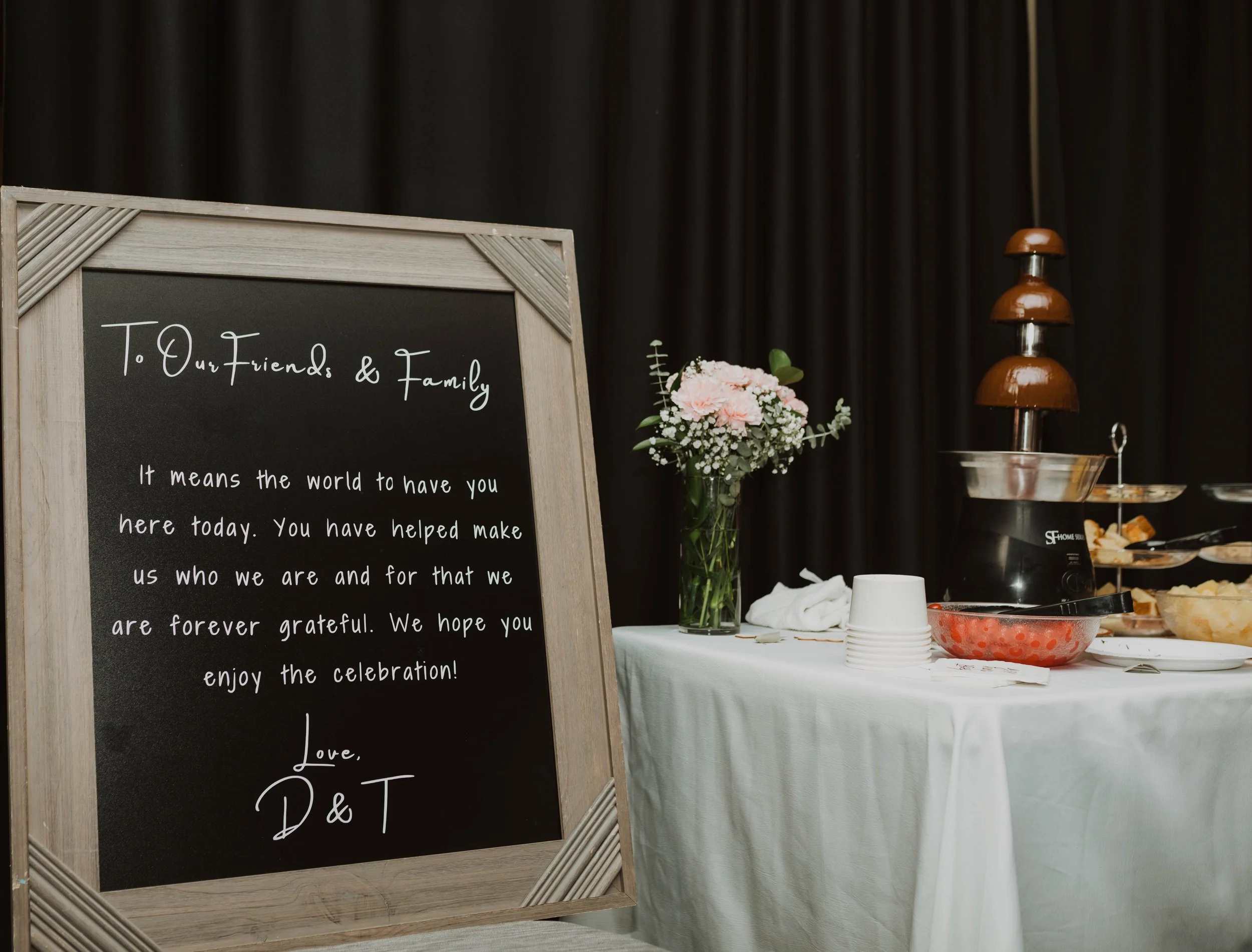 A blackboard sign with a wooden frame displaying a message for friends and family at a celebration, next to a table with a flower bouquet, a chocolate fountain, and serving bowls. Seattle, WA wedding photography.