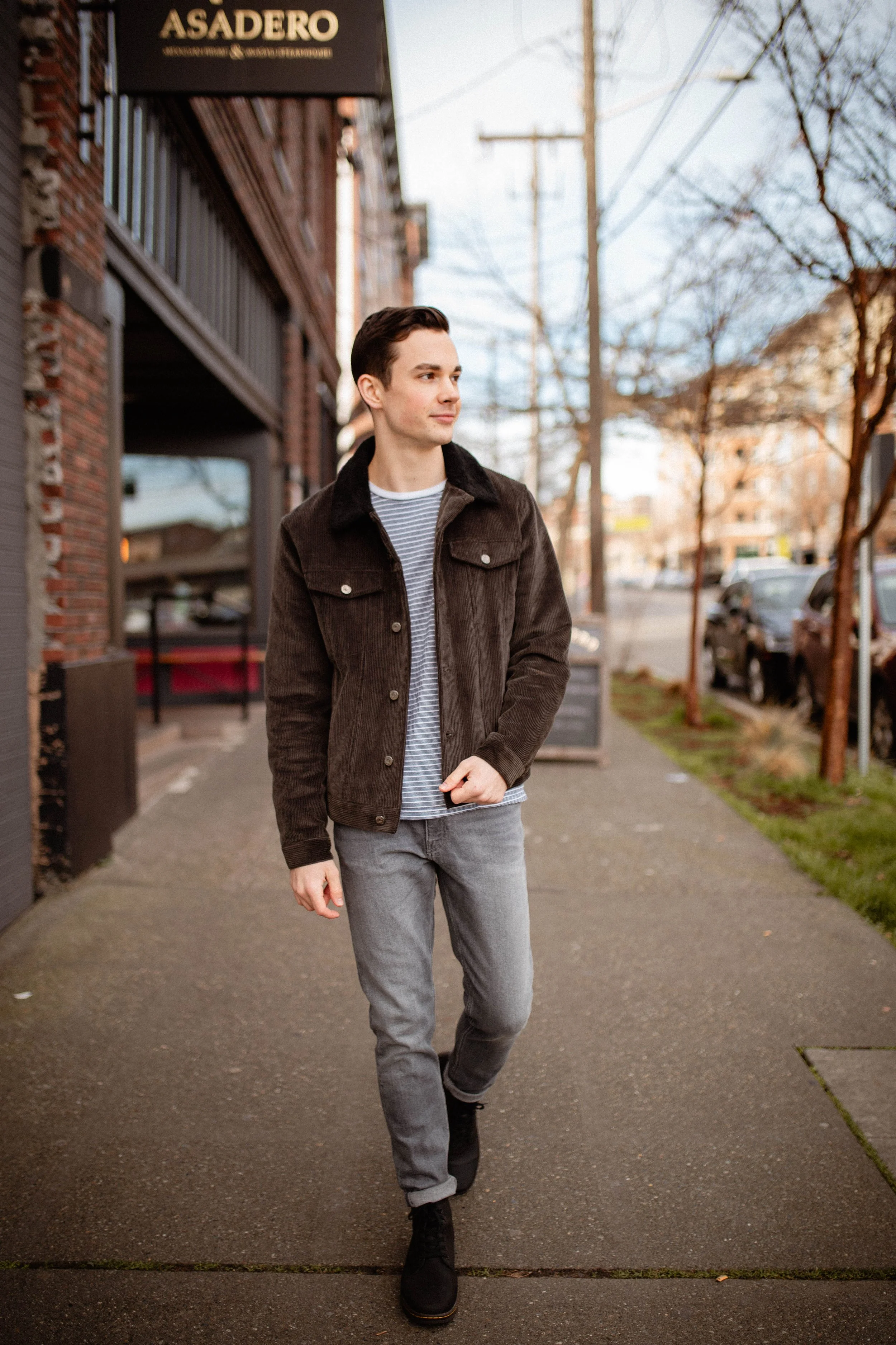 A young man walking on a sidewalk near a brick building with a sign that reads 'Asadero'. He is wearing a dark brown jacket, striped shirt, gray jeans, and black boots, and is looking to his left. Seattle professional head shot photography