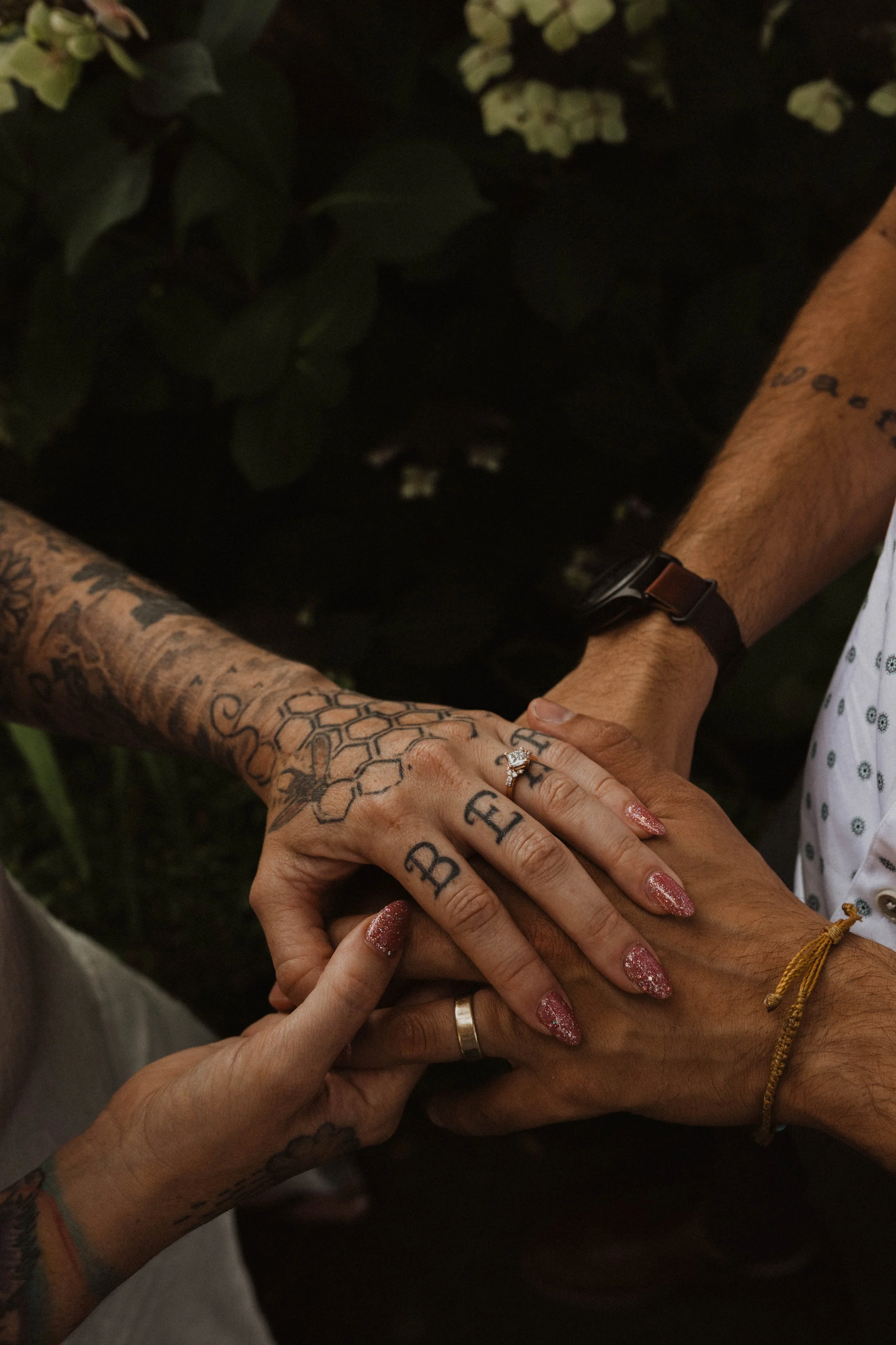 Close-up of three people stacking hands, showing tattoos, rings, and wristbands, with green foliage in the background. Seattle, WA wedding photography.