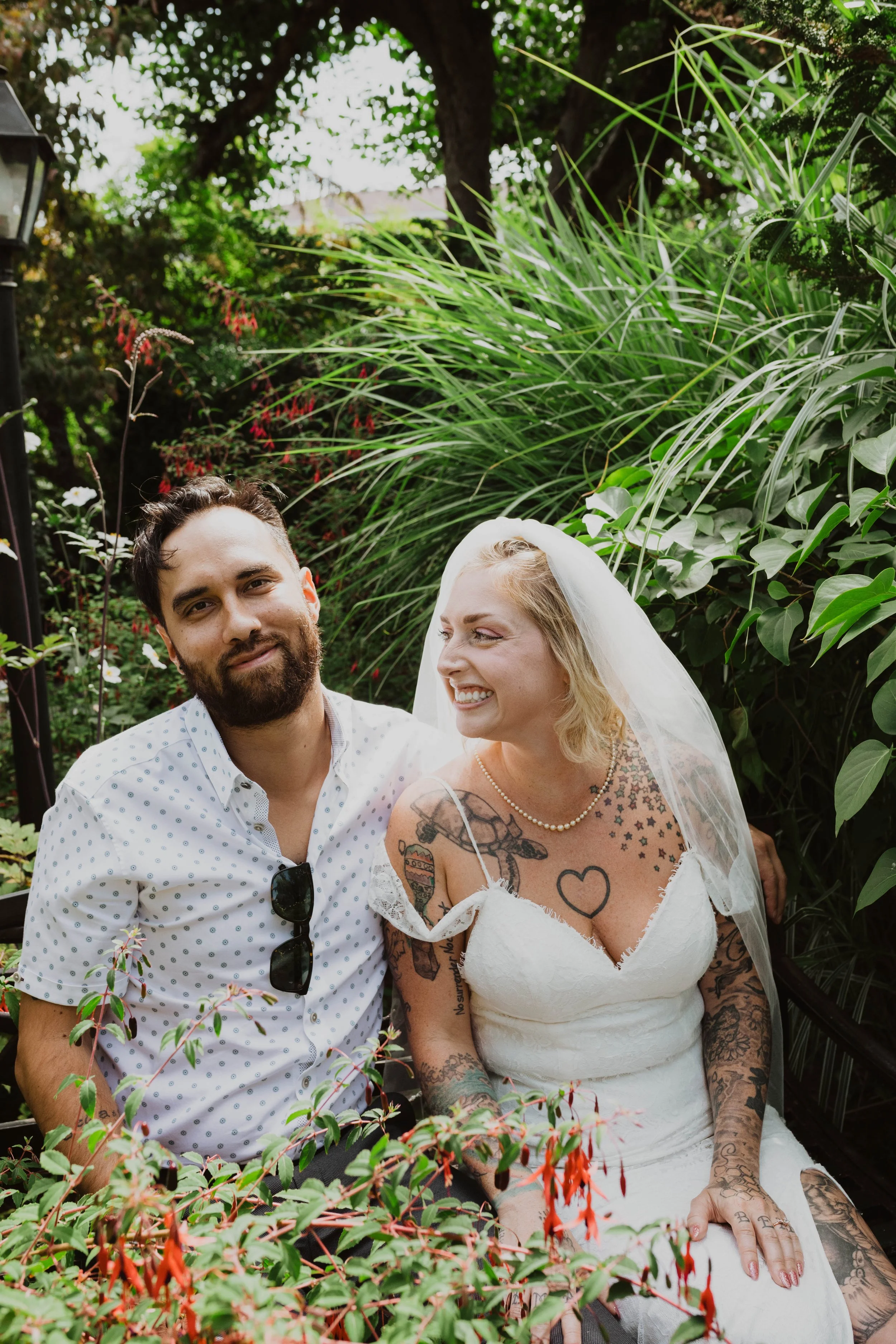 A smiling bride with tattoos sitting next to a bearded groom, surrounded by green plants and flowers in a garden setting. Seattle, WA wedding photography.