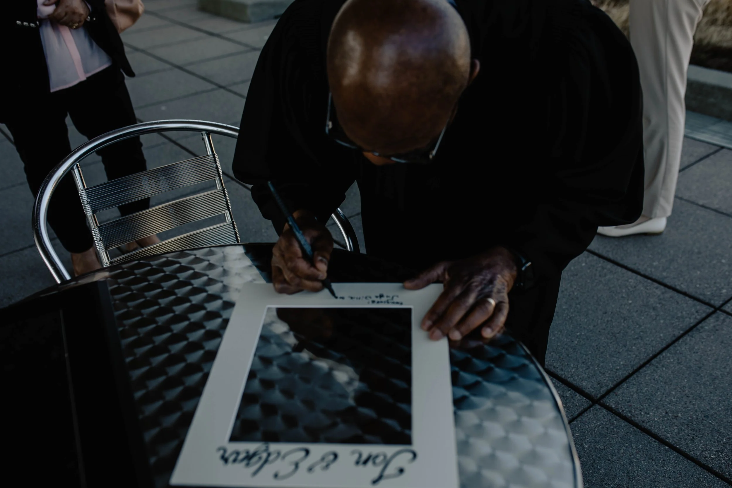 An elderly man with a bald head and glasses signs a framed photograph on a round metallic table outdoors at a public event, with other people standing nearby. Seattle Municipal Courthouse wedding photography.