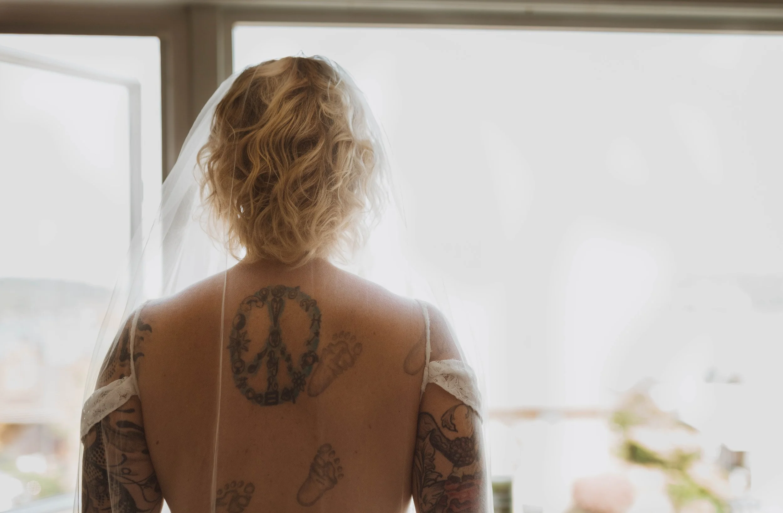 Back view of a woman with blonde curly hair, tattoos on her back and arms, wearing a white wedding dress and veil, standing near a window with bright daylight outside. Seattle, WA wedding photography.