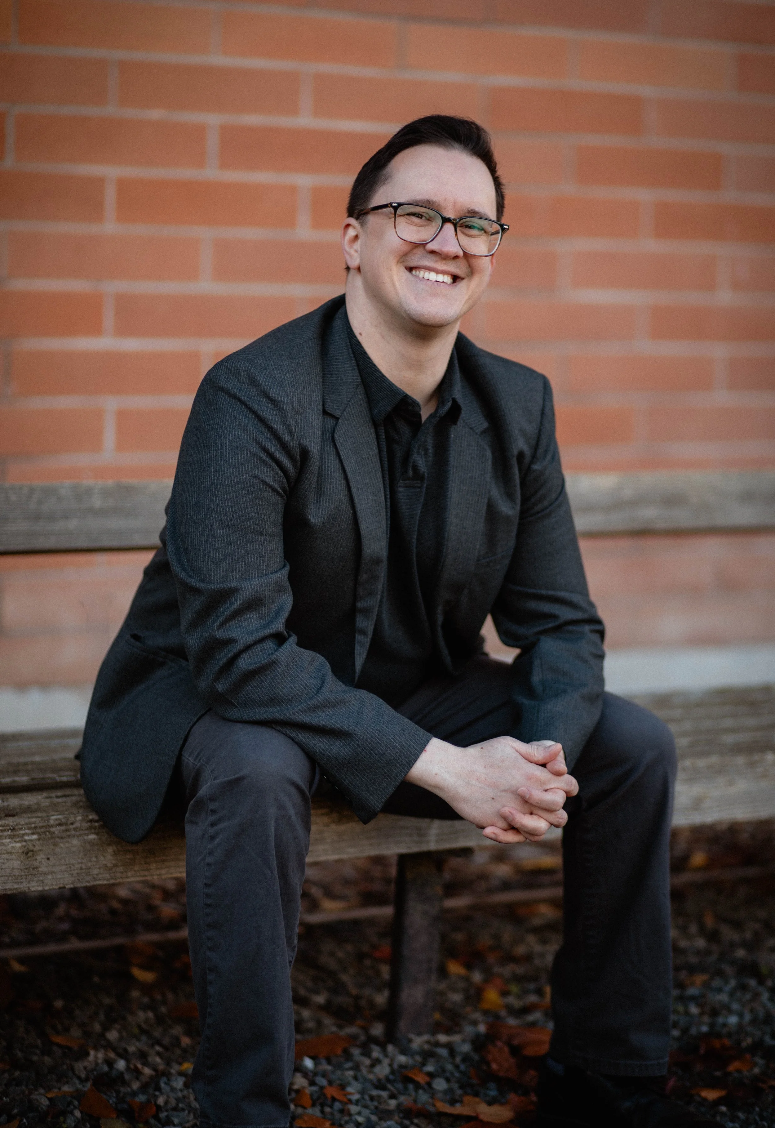 A man sitting on a wooden bench in front of a brick wall, wearing a black blazer and glasses, smiling at the camera. Seattle professional head shot photography