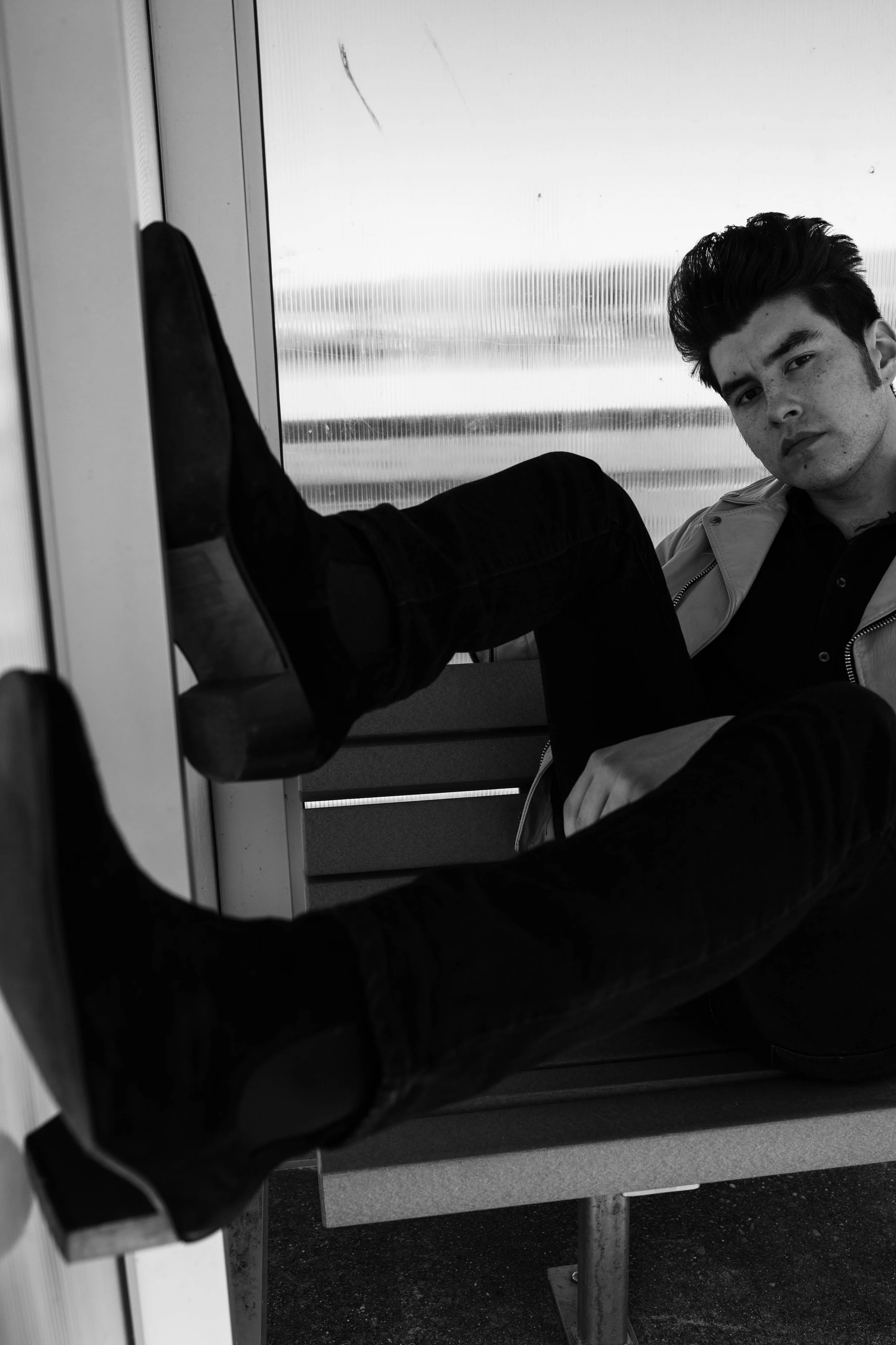 A young man sitting on a bench with his legs extended, wearing black shoes, dark pants, and a light-colored jacket. He is indoors near a window with blinds, and looks at the camera with a serious expression. Seattle professional head shot photography