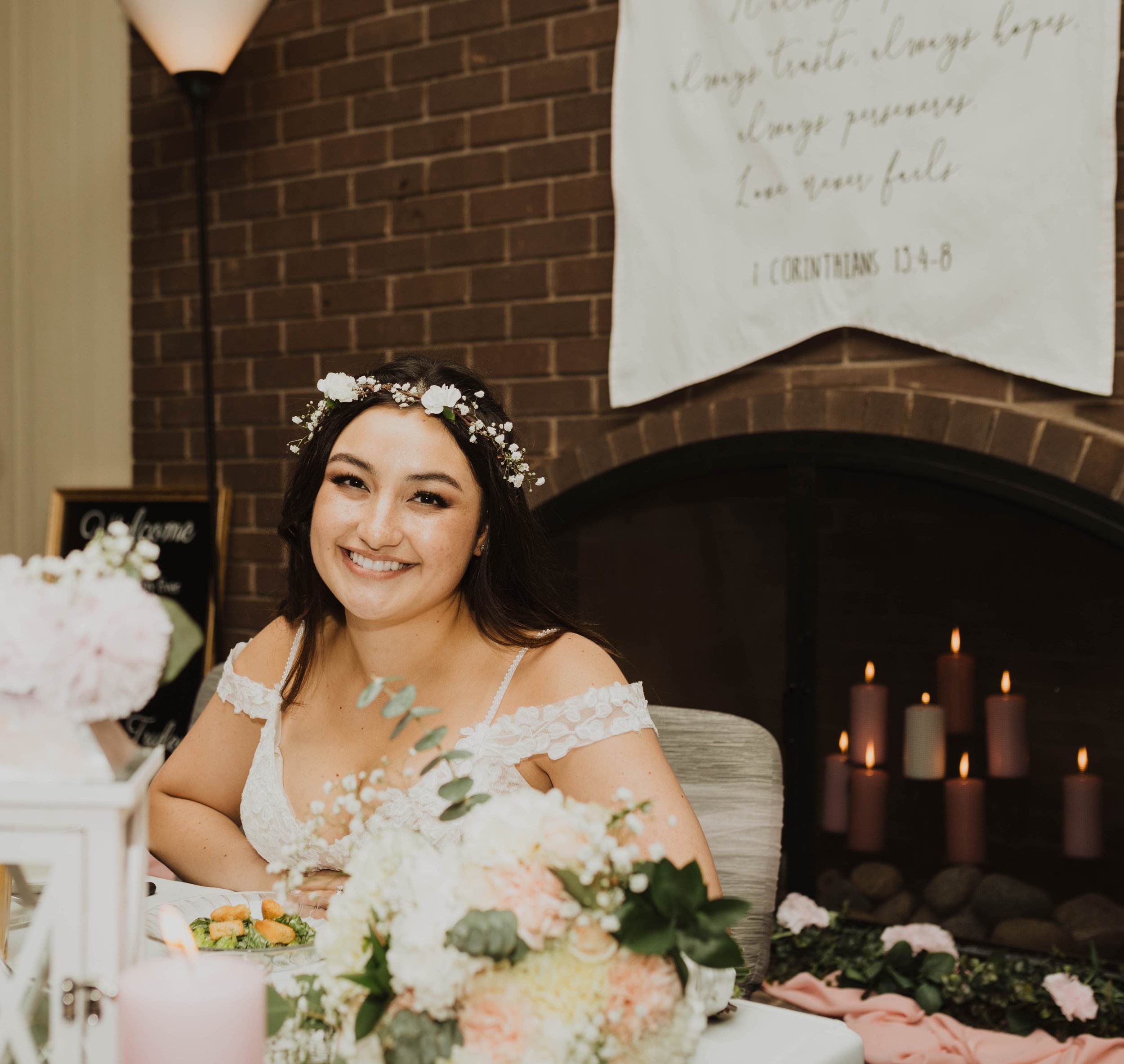 A woman smiling at a wedding reception, wearing a white dress and a floral headband. There are candles and floral decorations on the table, and a brick fireplace with lit candles in the background. Seattle, WA wedding photography.