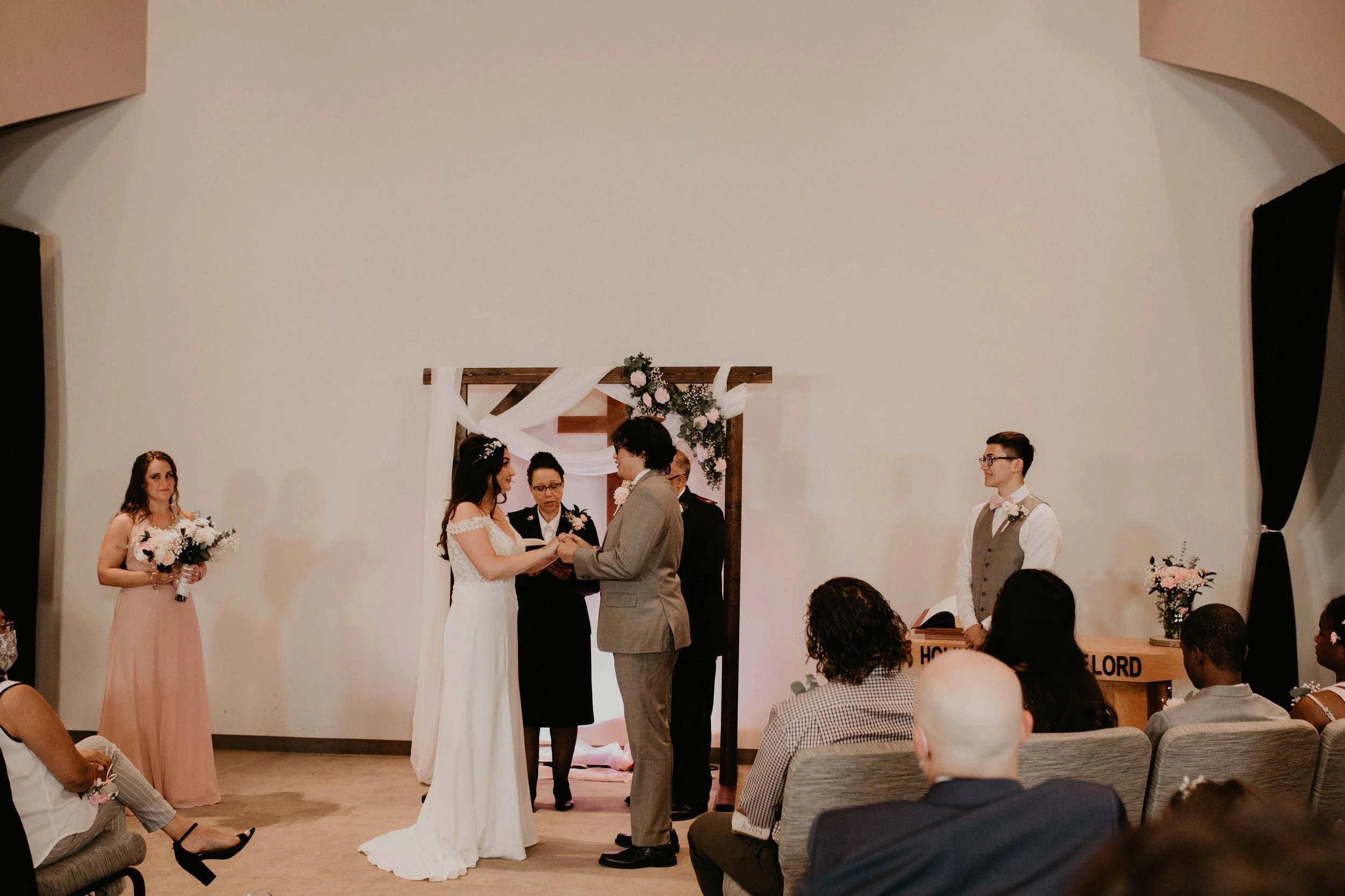A wedding ceremony with a bride and groom holding hands at the altar, officiant gesturing, bridesmaid holding flowers on the left, and groomsman standing on the right, with guests seated watching. Seattle, WA wedding photography.