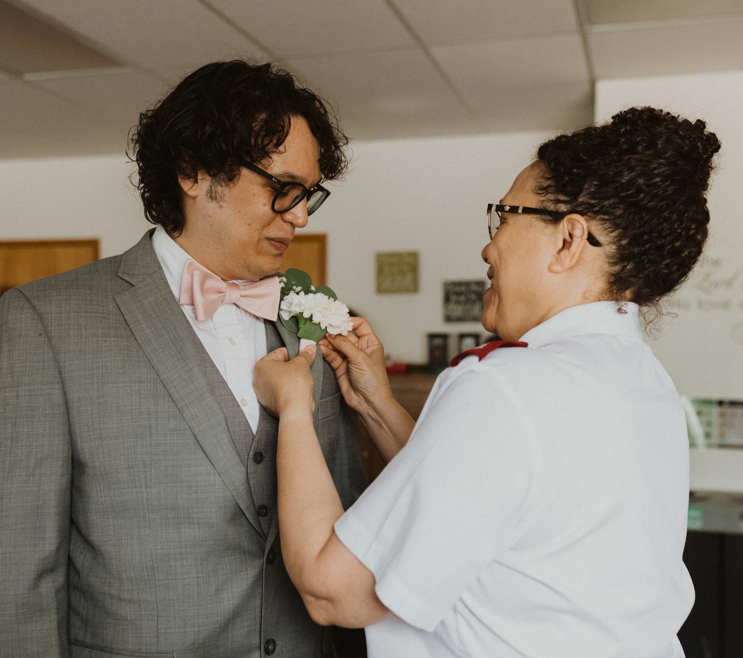 A person in a suit and pink bow tie gets a white boutonniere pinned on their lapel by a woman in a white uniform with red shoulder accents. Seattle, WA wedding photography.