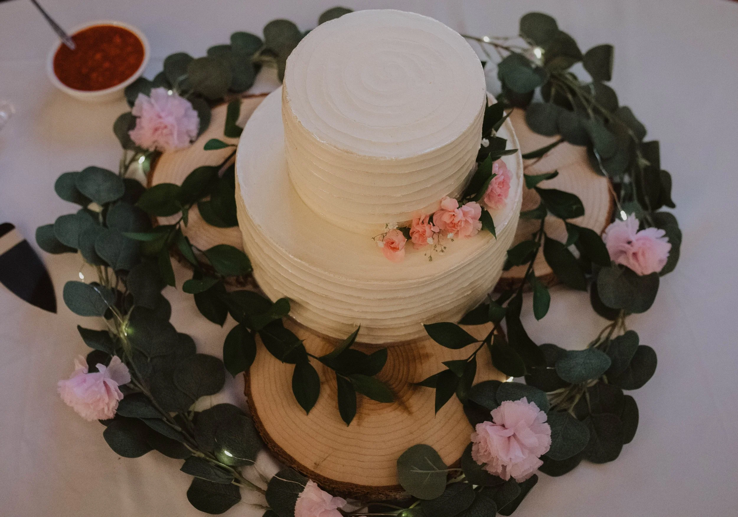 A white, tiered wedding cake with pink flowers on a wooden slab surrounded by green leaves and pink flowers, on a table with a small bowl of red sauce in the background. Seattle, WA wedding photography.
