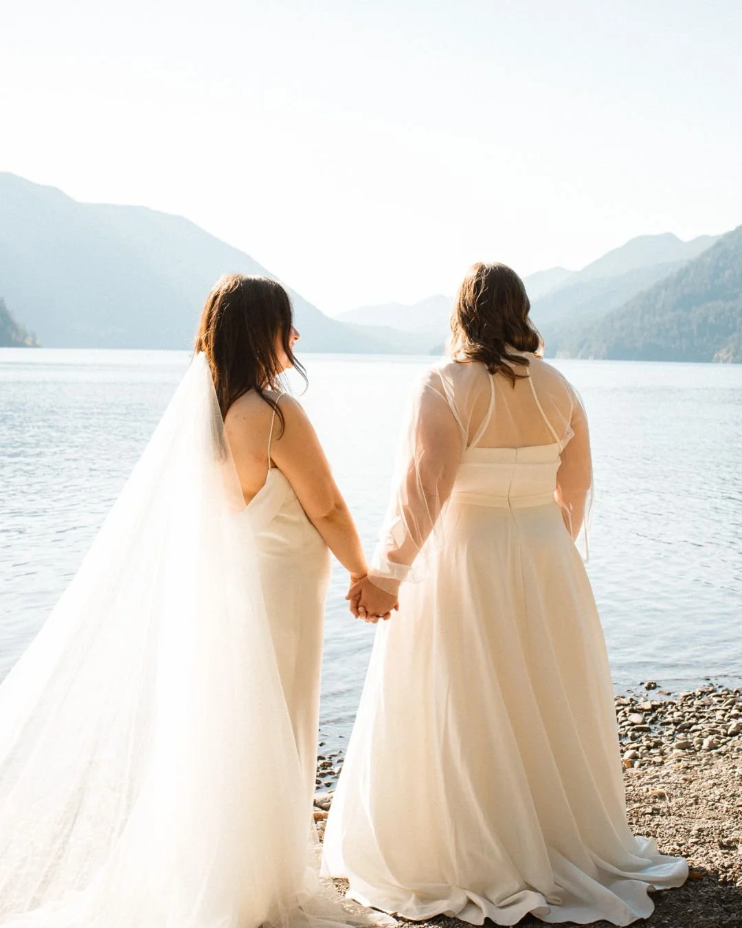 Two women holding hands and smiling at each other on a rocky lakeside, with mountains and a calm lake in the background, dressed in white wedding gowns. Wedding photography at Lake Crescent in Port Angeles, WA