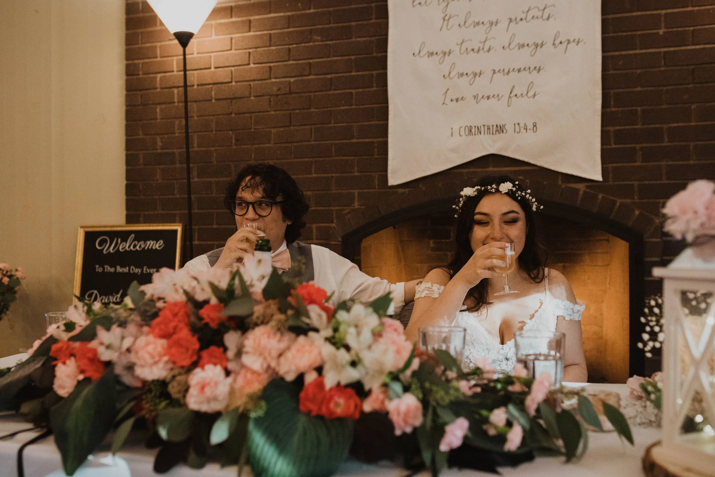 A bride and groom sitting at a wedding reception table, drinking from glasses, with a floral centerpiece in front of them and a brick fireplace in the background. Seattle, WA wedding photography.