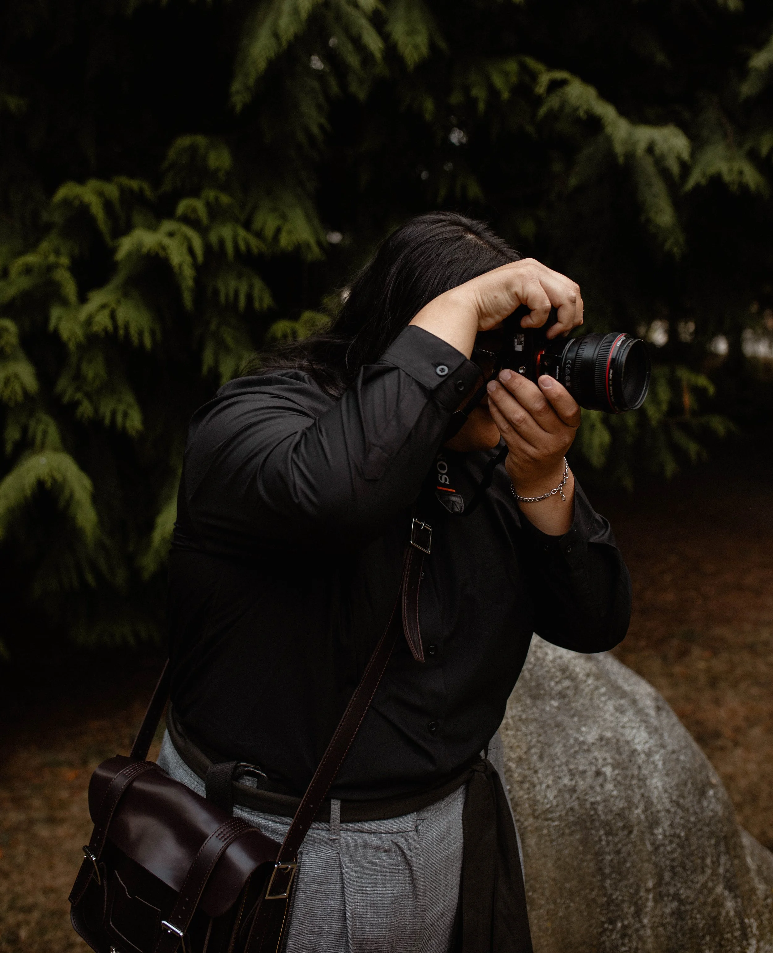 Person with dark hair in black shirt and gray pants taking a photograph with a camera outdoors, with trees and rocks in the background. Seattle professional head shot photography