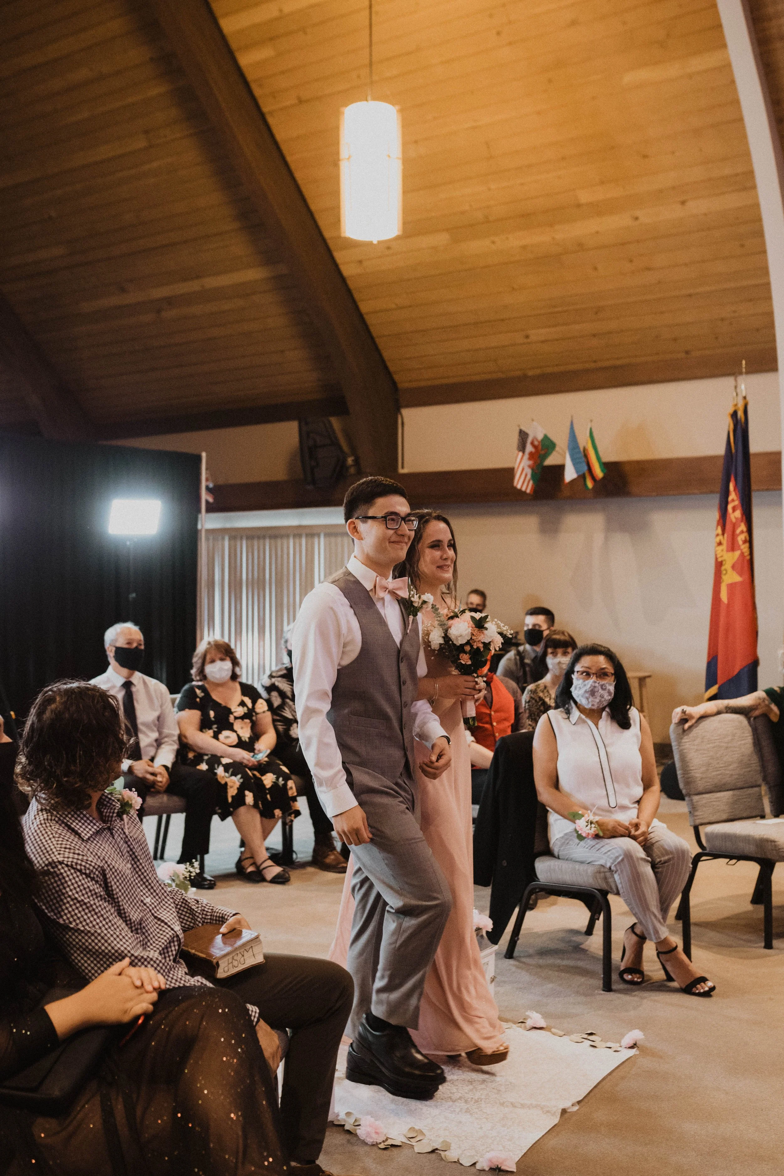 A couple walking down the aisle with guests seated on both sides during a wedding ceremony in a wooden church, with some guests wearing face masks. Seattle, WA wedding photography.