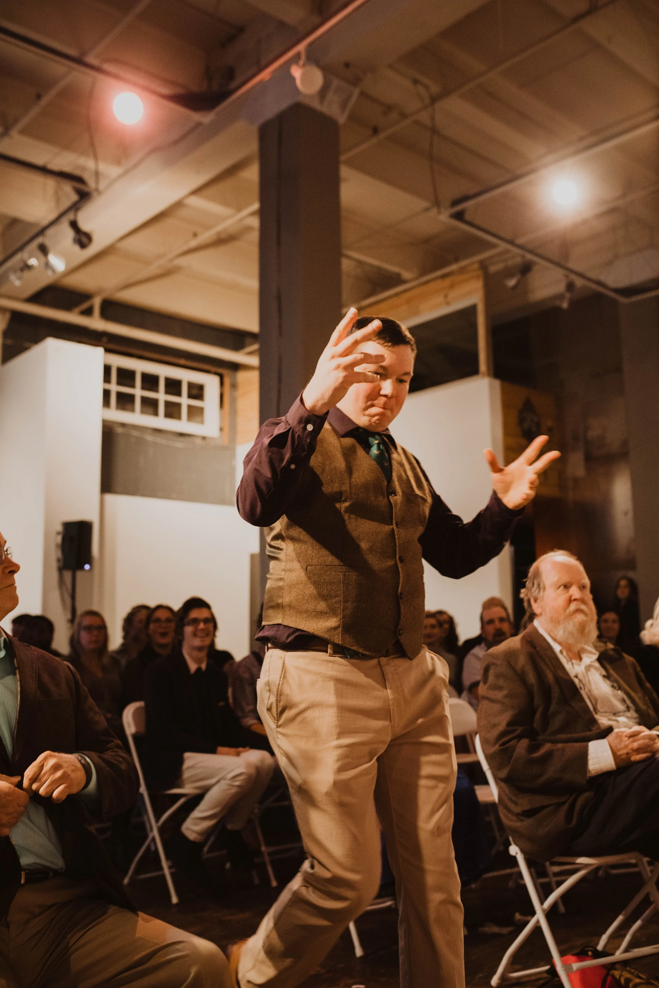A young man stands in front of an audience with a lively expression, gesturing with his hands, in a room with a high ceiling, wooden beams, and audience members seated on chairs. Pioneer Square, Seattle, WA wedding photography.