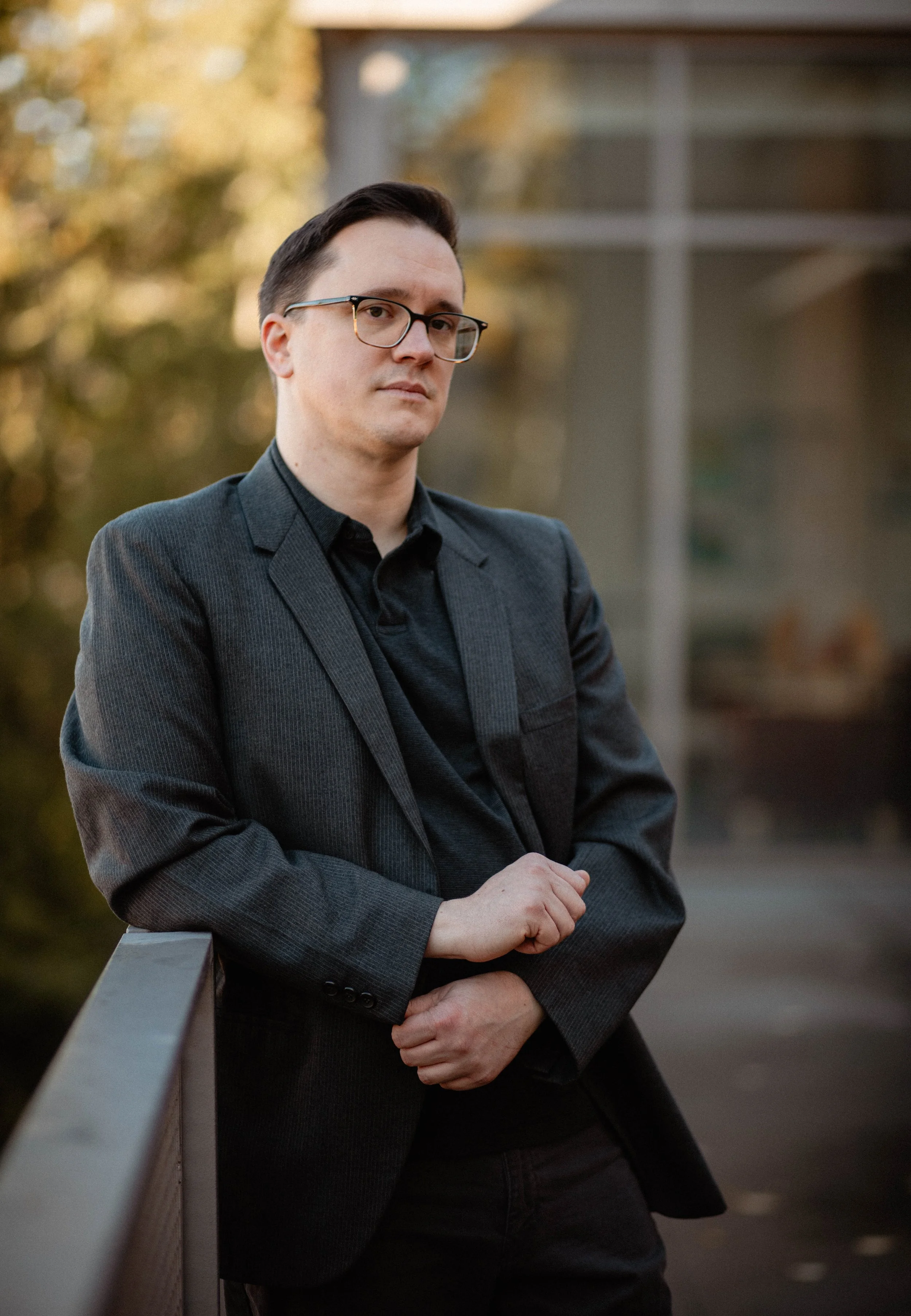 A man with dark hair, glasses, and a black suit jacket standing outdoors near a railing, with trees and a building in the background during sunset. Seattle professional head shot photography