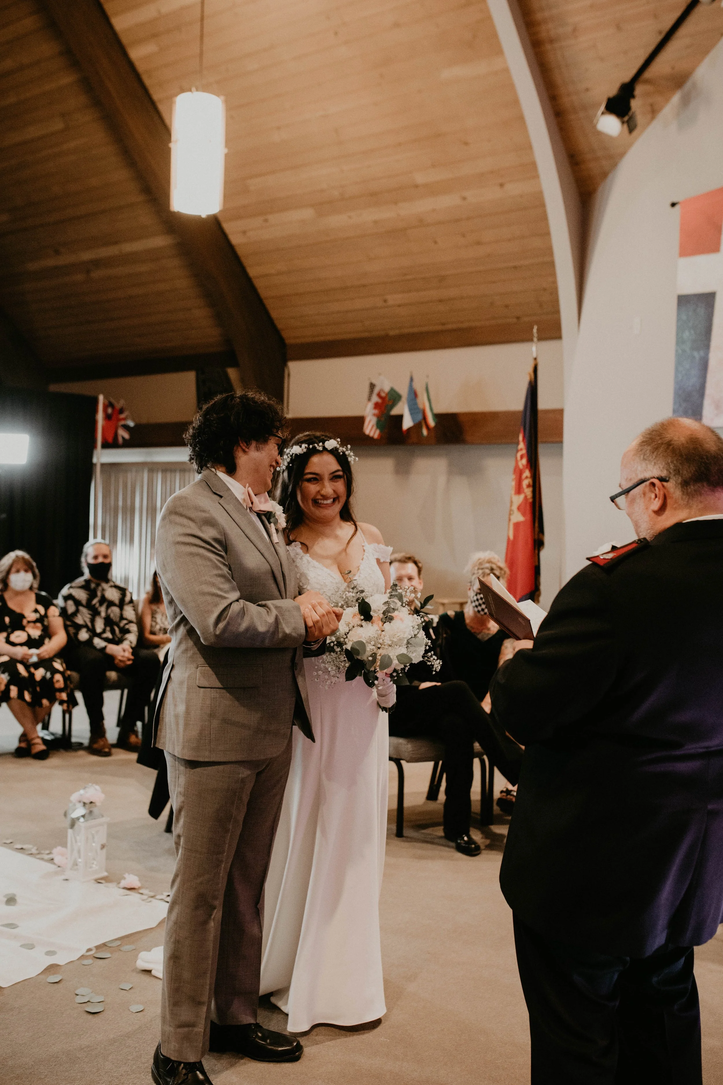 A wedding ceremony with a couple exchanging vows in front of an officiant, with seated guests in the background, some wearing masks, and international flags displayed behind them. Seattle, WA wedding photography.