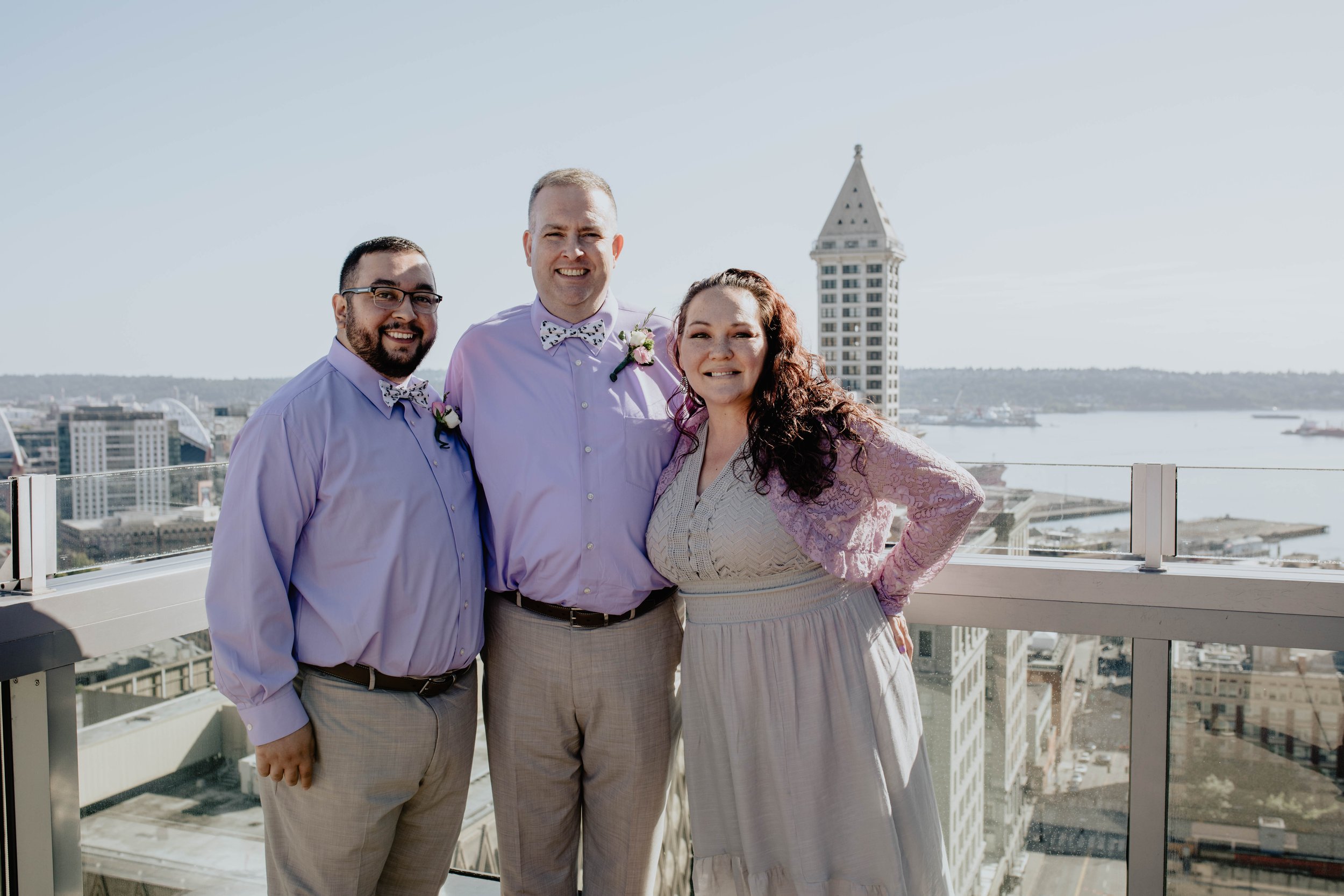 Three people standing on a rooftop with a city skyline and water in the background, dressed in formal attire for a celebration or event. Seattle Municipal Courthouse wedding photography.