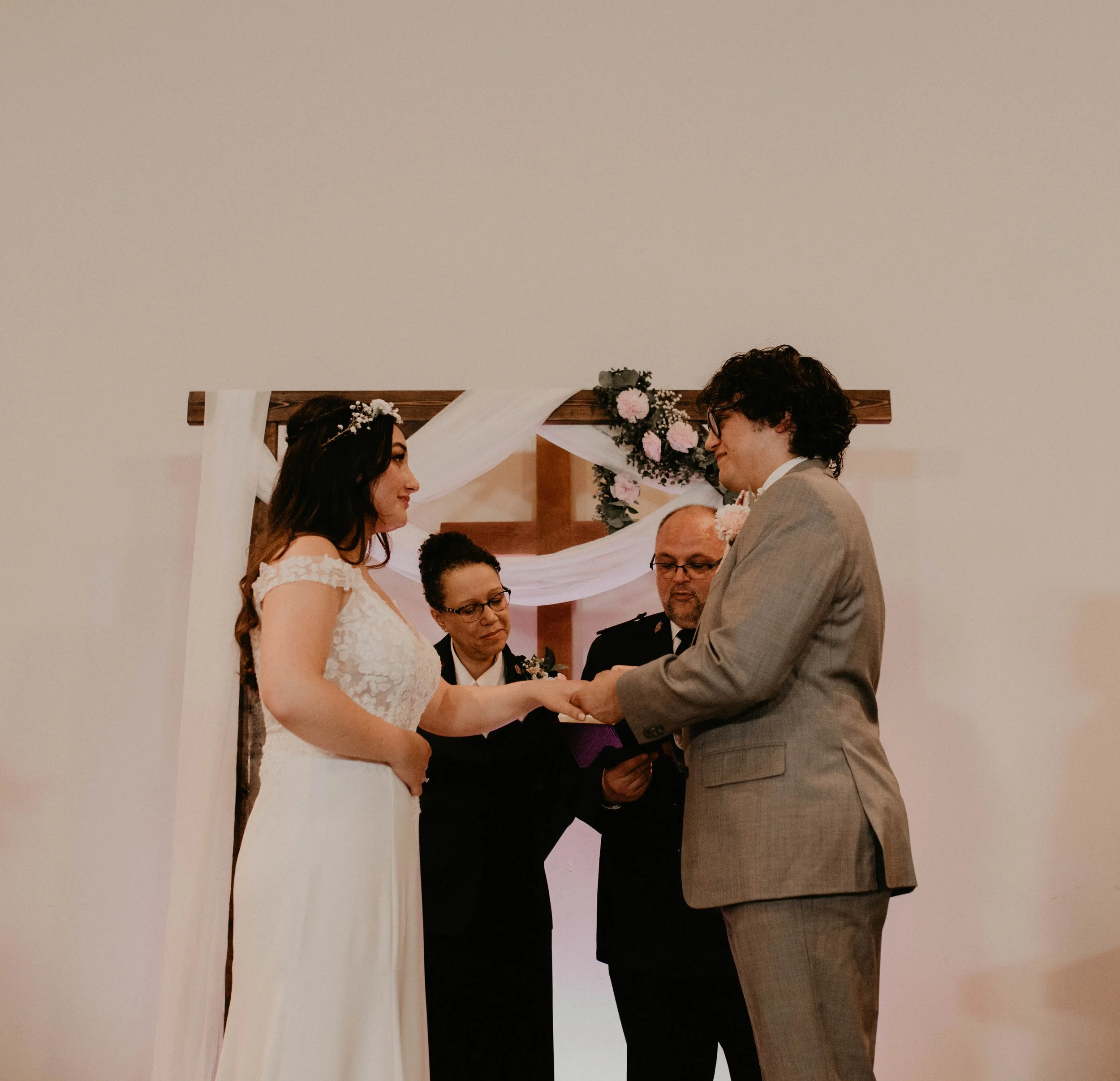 A couple exchanging rings during a wedding ceremony, with officiants witnessing them, in front of a wooden arch decorated with white fabric and pink flowers. Seattle, WA wedding photography.