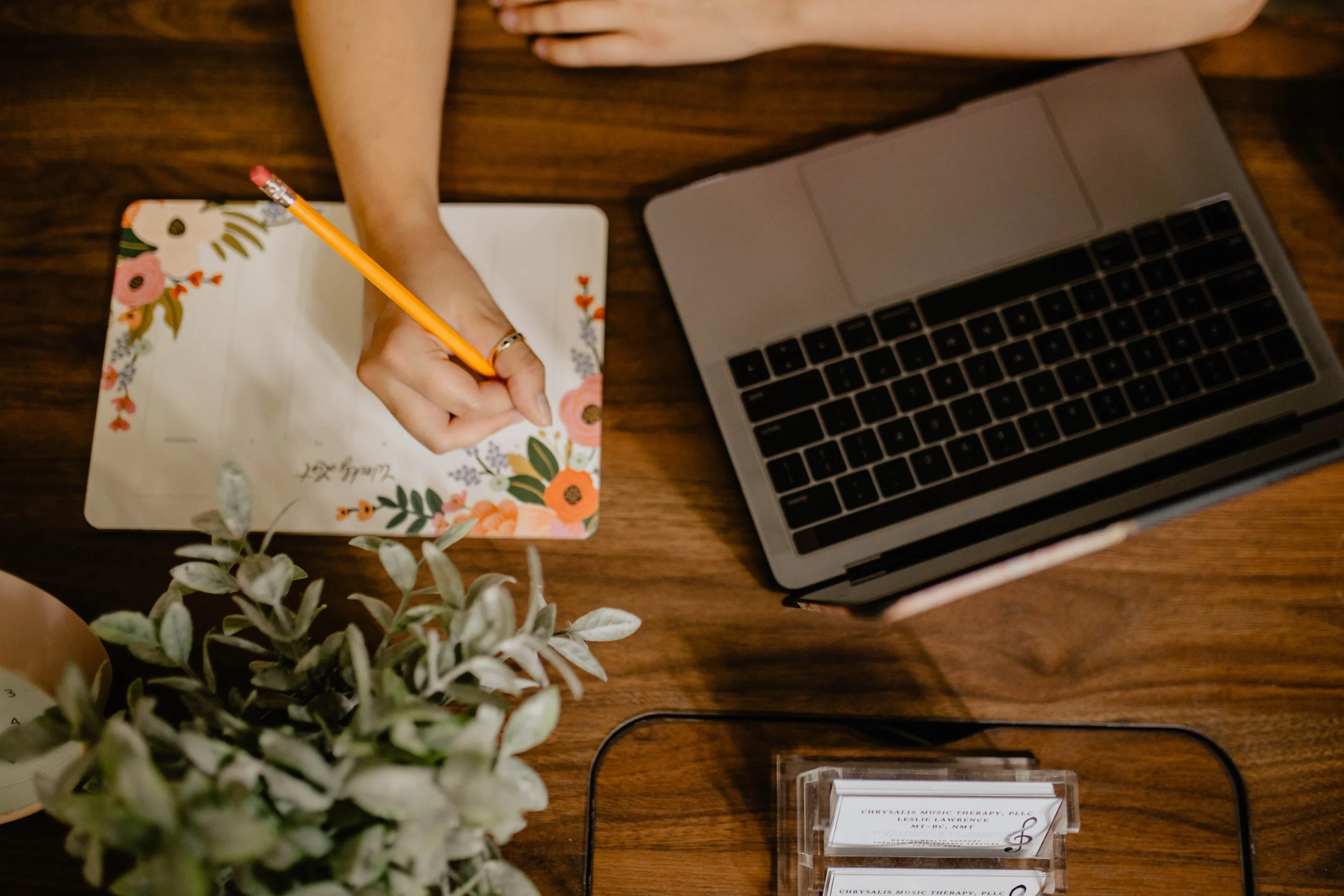 A person is writing on a floral decorated planner with a pencil, next to an open laptop on a wooden table, with a potted plant and a business card holder nearby. Seattle professional head shot photography
