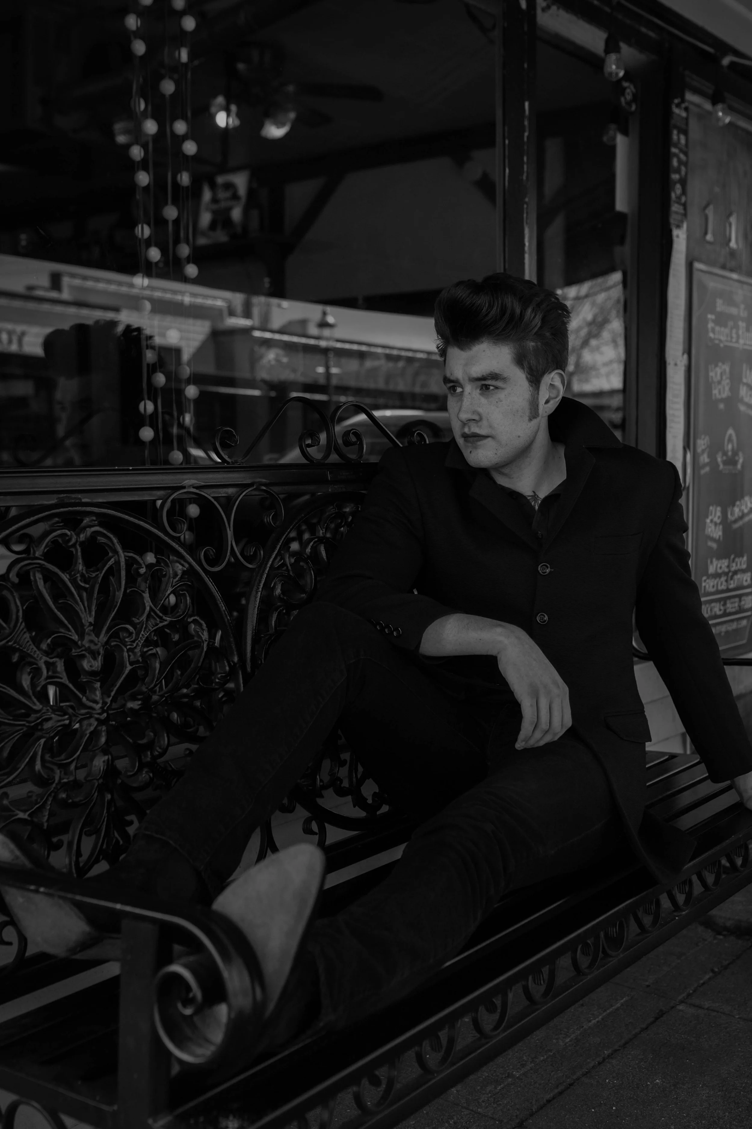 A young man with styled hair, sitting on a decorative iron bench outside a storefront, looking to his right with a serious expression, in black and white. Seattle professional head shot photography