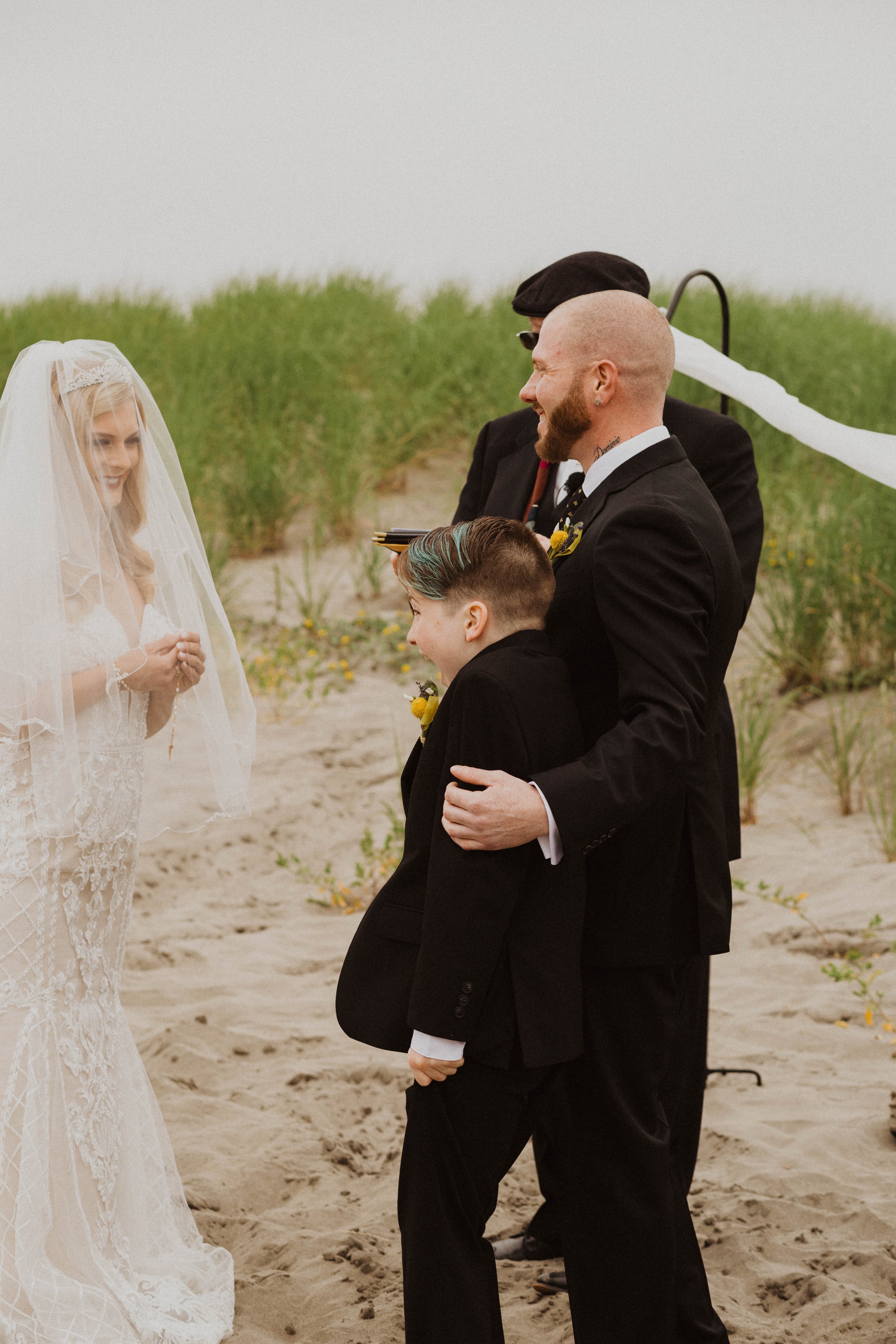 A beach wedding ceremony with a bride in a lace wedding dress and veil, and groom in a black suit, exchanging vows with two boys in suits, one smiling and the other looking down, amid sandy terrain and green beach grass. Long Beach, WA wedding photog