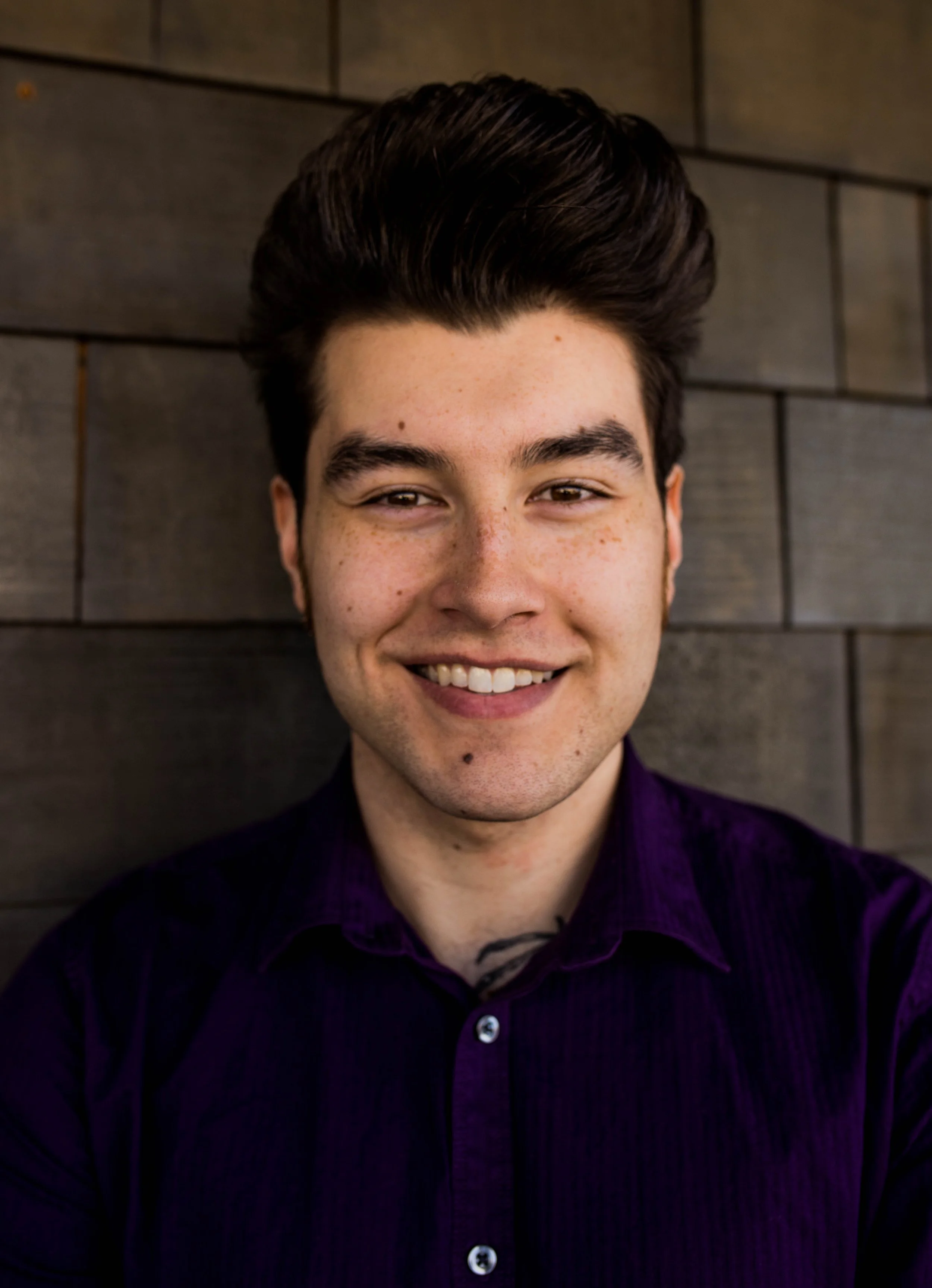 A smiling young man with dark, neatly styled hair, wearing a dark purple shirt, standing in front of a wooden wall. Seattle professional head shot photography