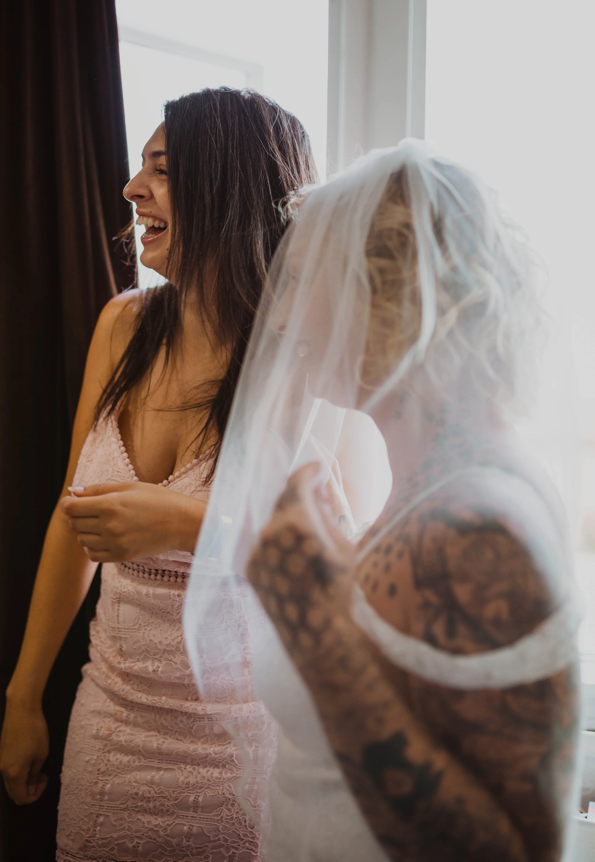 Two women, one in a pink lace dress and the other in a veil with tattoos on her arm, laughing and sharing a joyful moment near a window. Seattle, WA wedding photography.