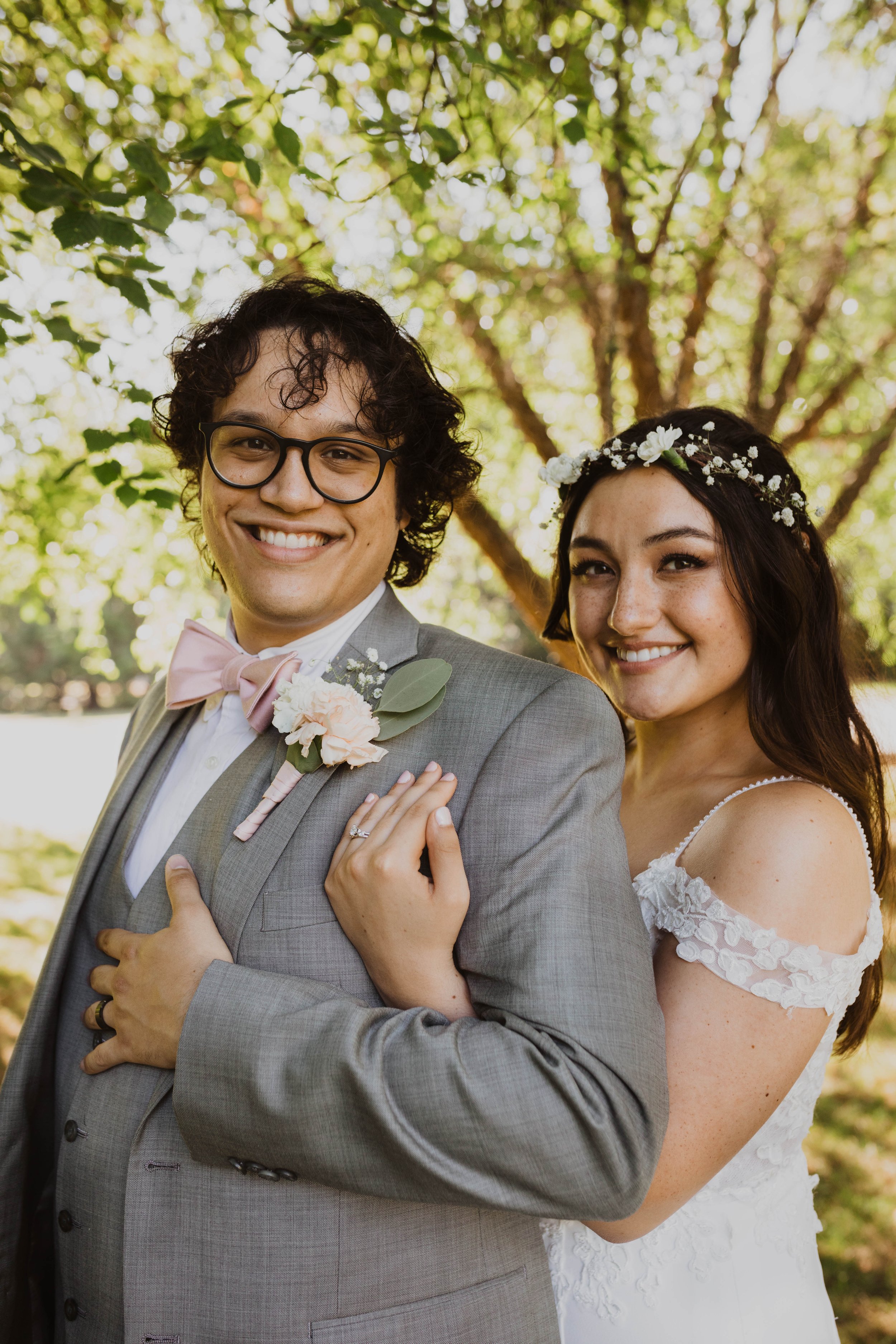 A newlywed couple smiling outdoors, with the groom in glasses, a grey suit, floral boutonniere, and pink bow tie, and the bride wearing a white lace off-shoulder wedding dress and a floral headband. Seattle, WA wedding photography.