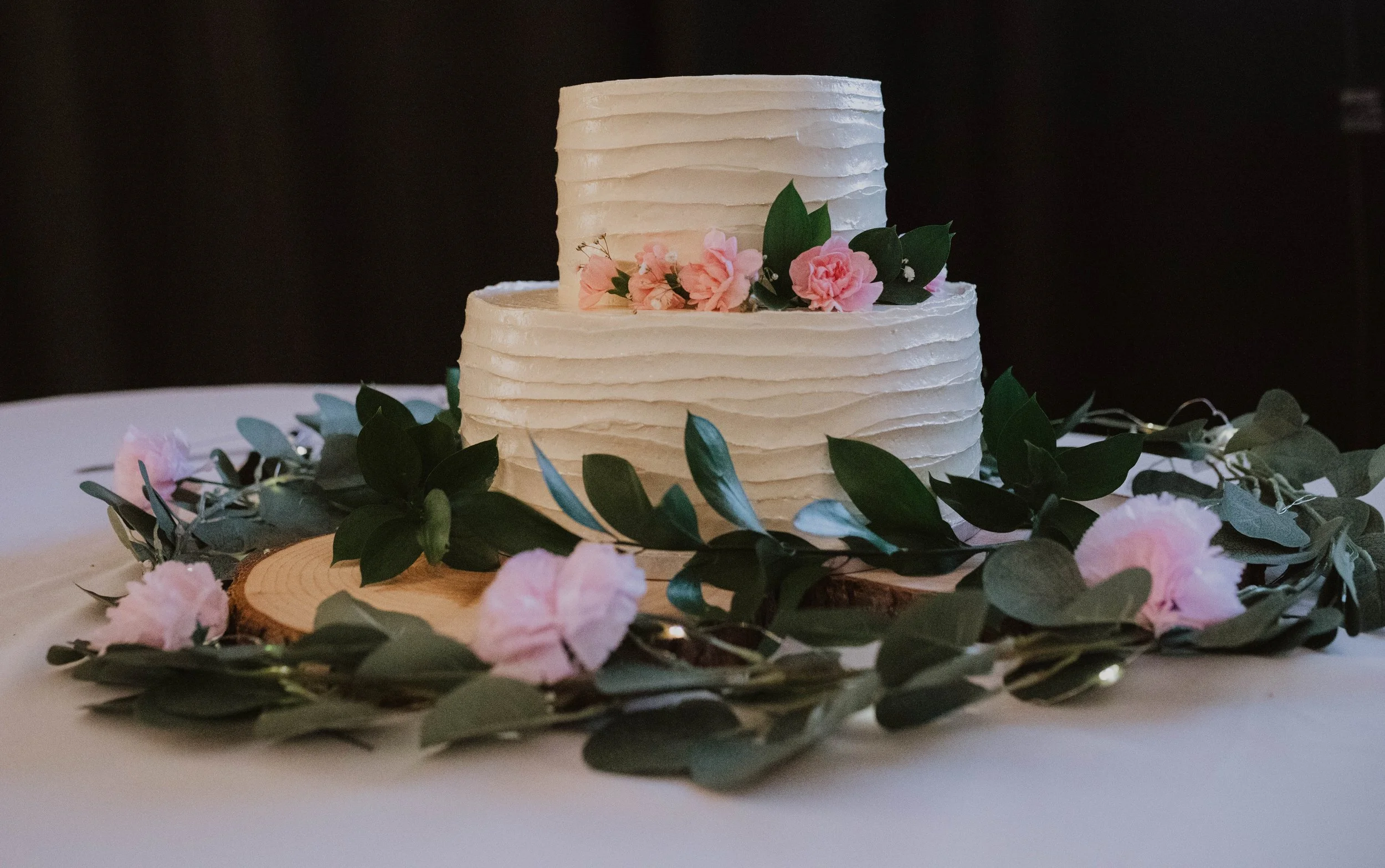 Two-tiered white wedding cake with textured frosting, decorated with pink flowers and green leaves, on a wooden slab surrounded by pink flowers and greenery. Seattle, WA wedding photography.