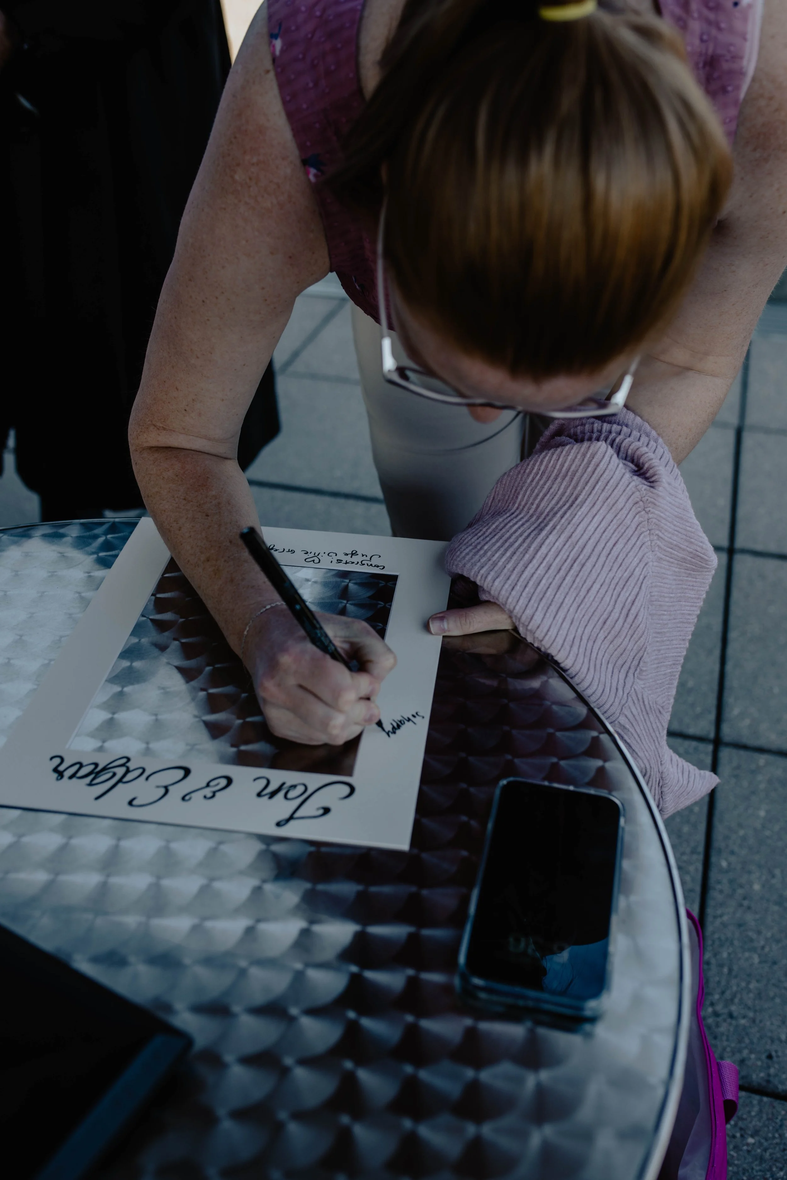 A woman with red hair signing a poster on a metal table, with a child's arm holding the poster. There is a smartphone on the table. Seattle Municipal Courthouse wedding photography.
