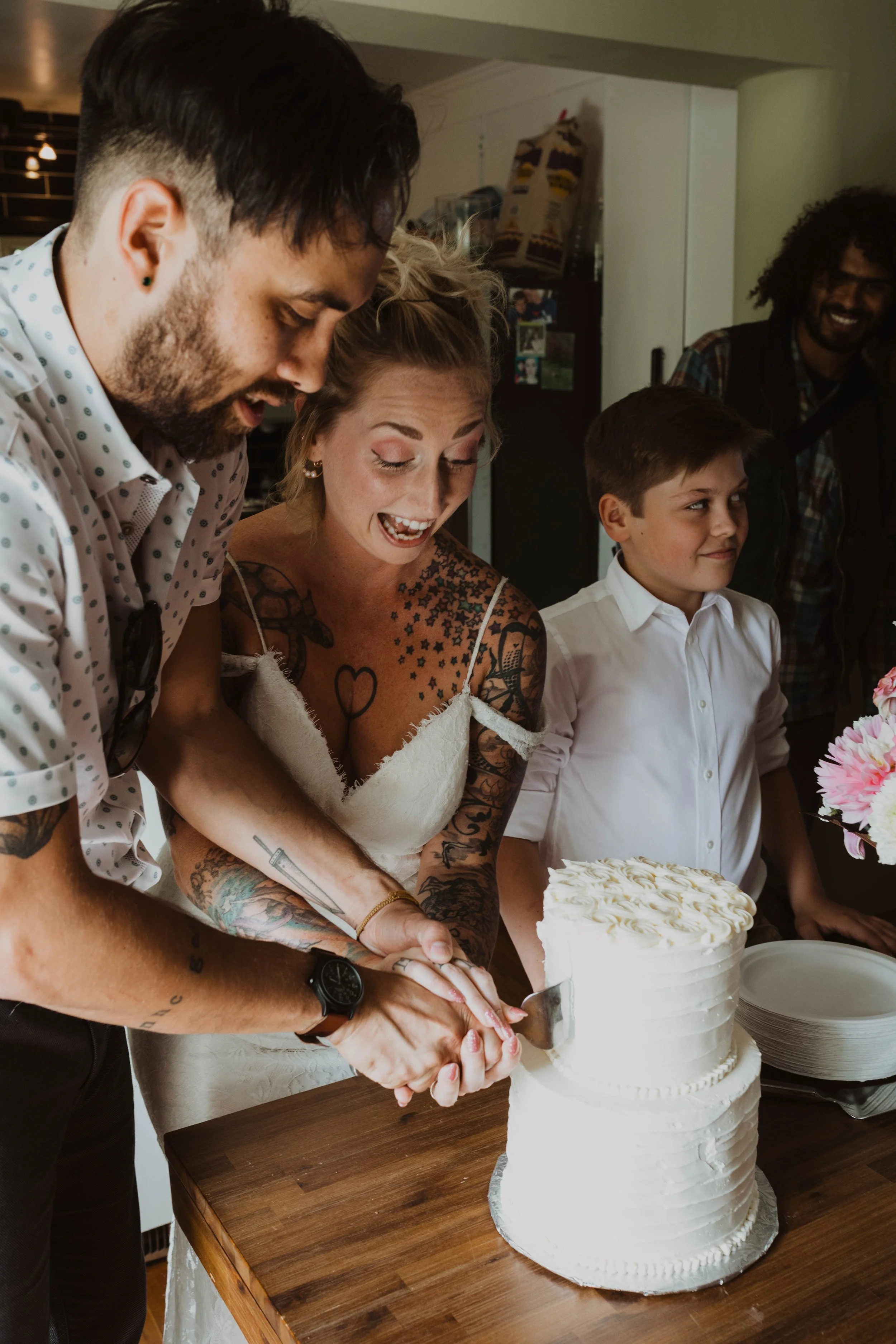 A couple is cutting a wedding cake at a celebration. The bride has tattoos and is smiling, while the groom is holding the knife. A young boy and two other people are standing nearby, smiling and watching the cake-cutting. Seattle, WA wedding photogra