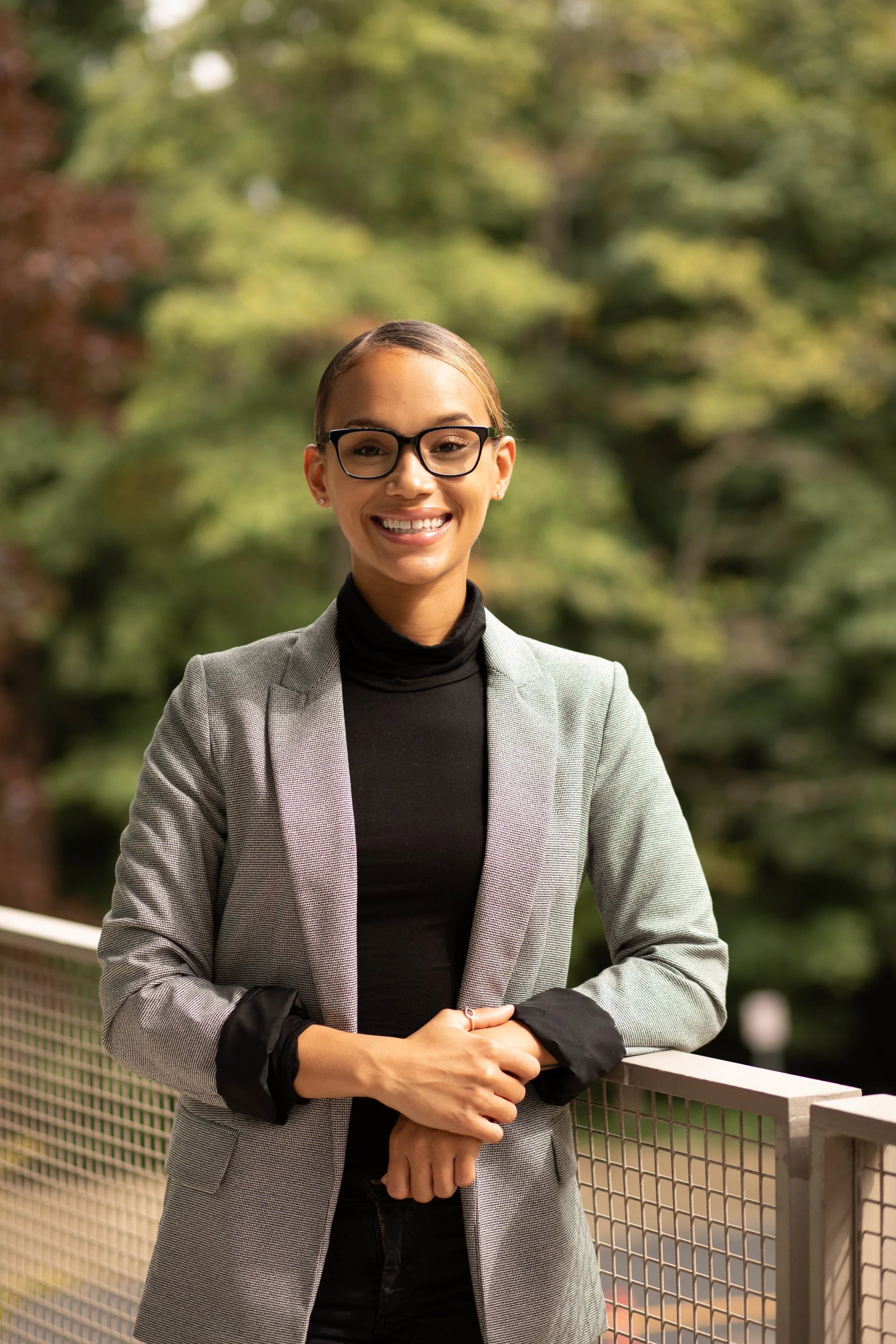 A woman with glasses, wearing a gray blazer and black turtleneck, standing outdoors near a railing with green trees in the background. Seattle professional head shot photography
