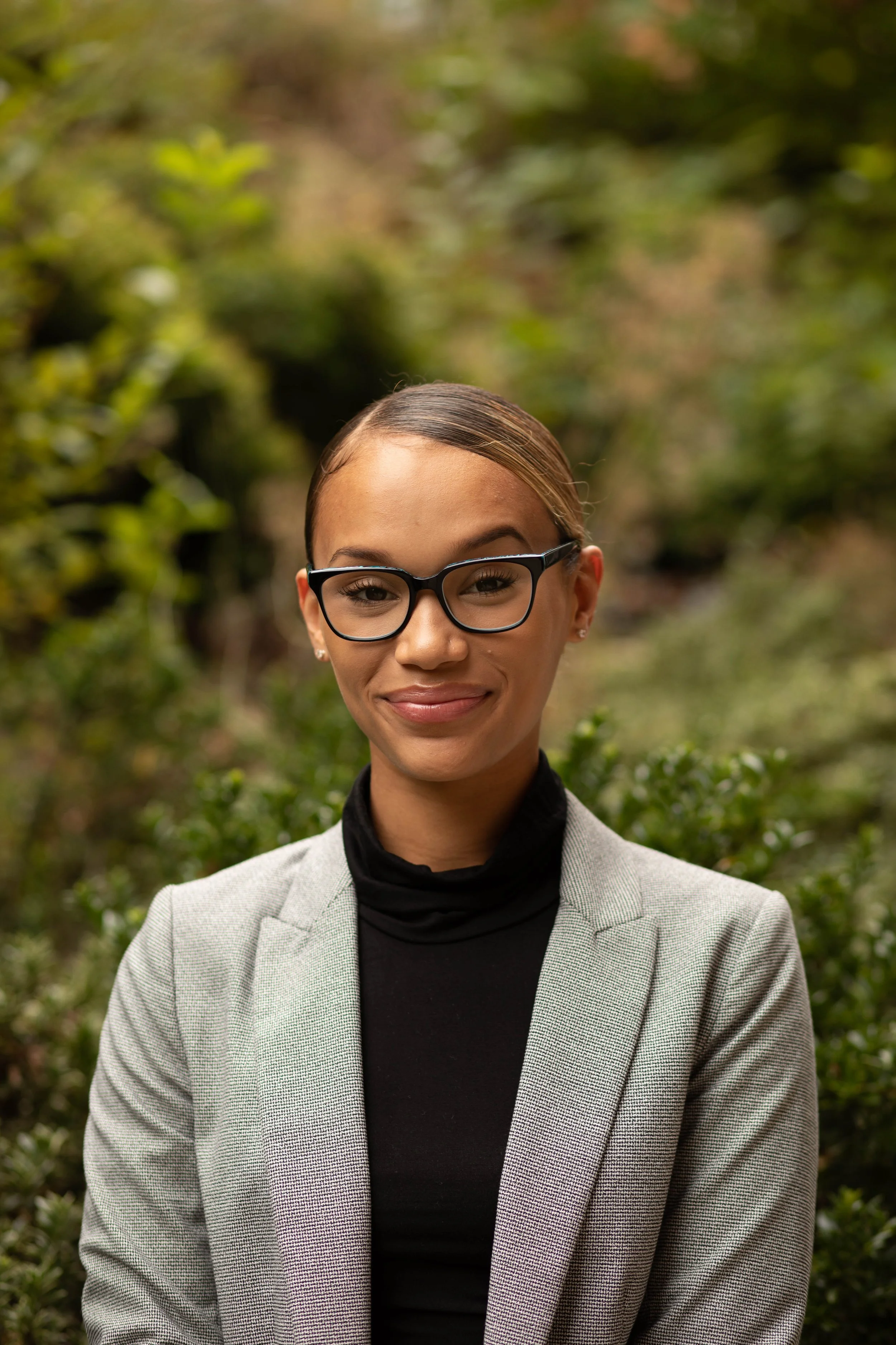 A woman wearing glasses and a gray blazer, standing outdoors with a blurred green background. Seattle professional head shot photography