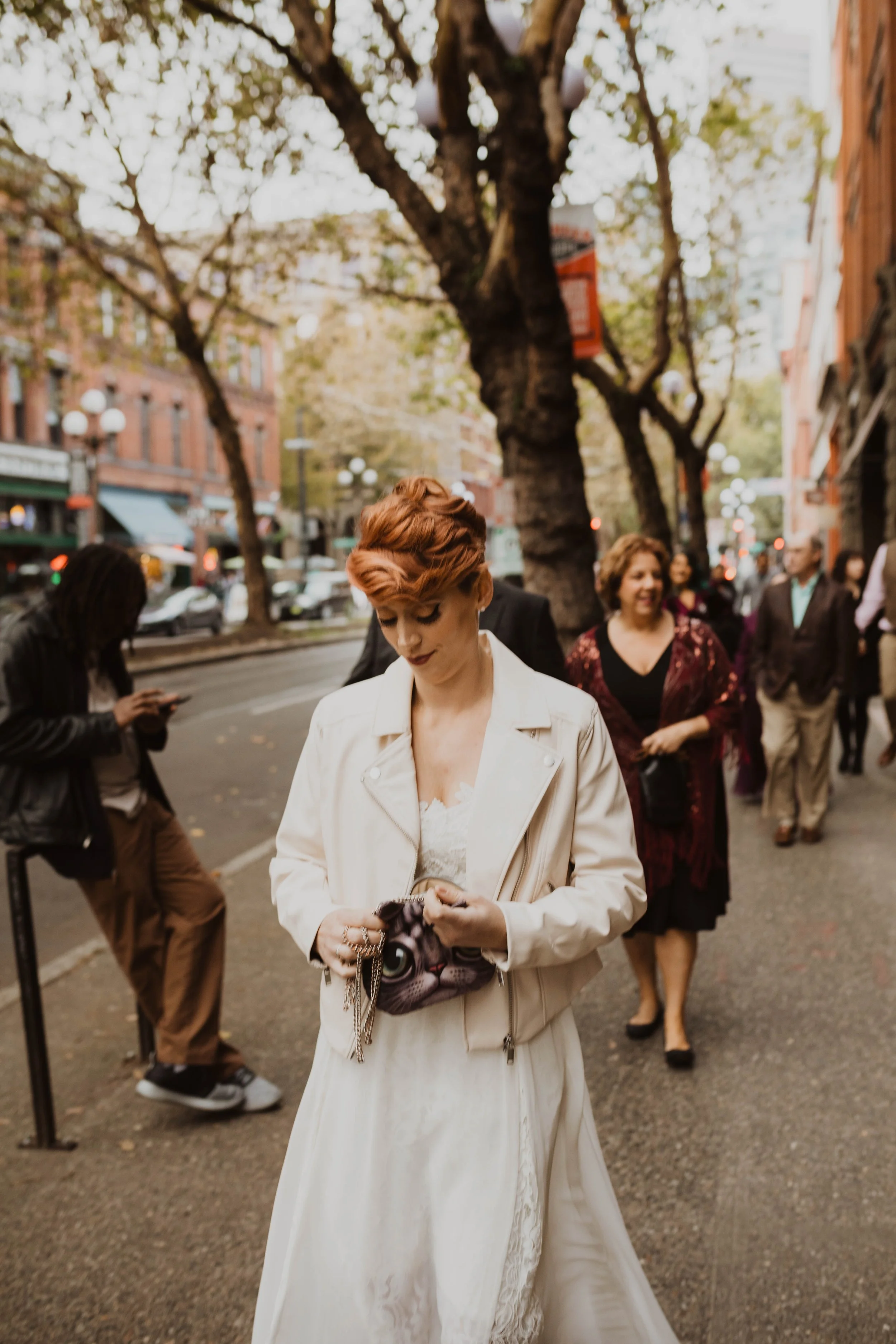 A woman with short red hair in a white dress and leather jacket standing on a city street, looking at her purse, with other people walking behind her under a large tree. Pioneer Square, Seattle, WA wedding photography.