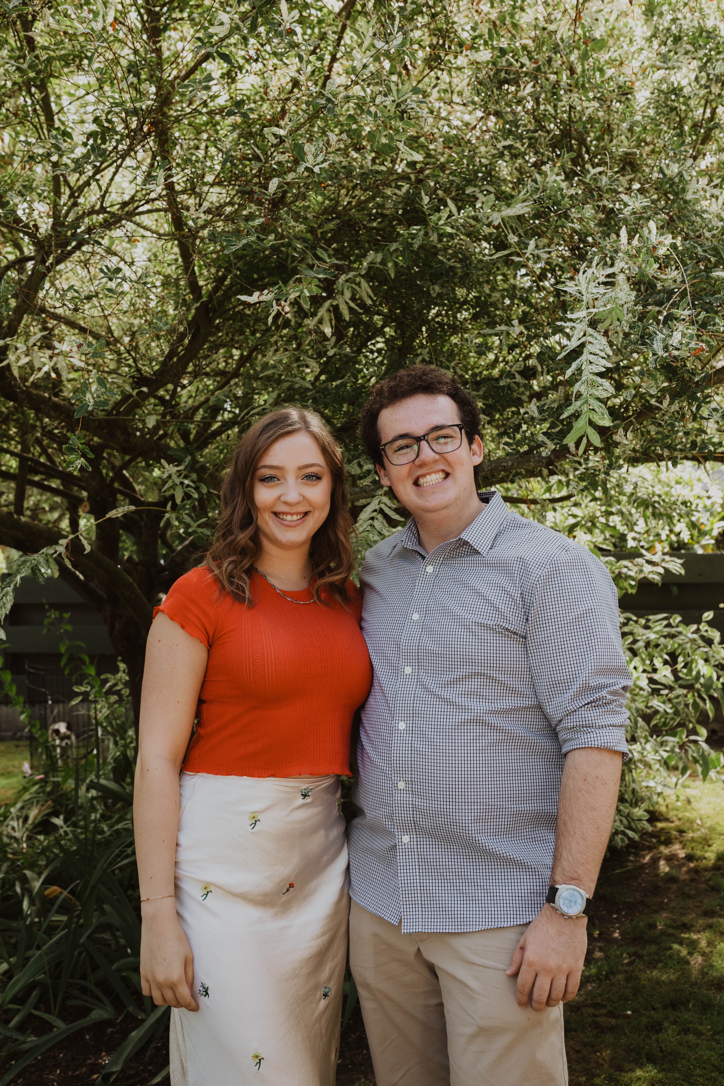 A young couple standing arm-in-arm outdoors beneath a tree, smiling at the camera. Seattle, WA wedding photography.