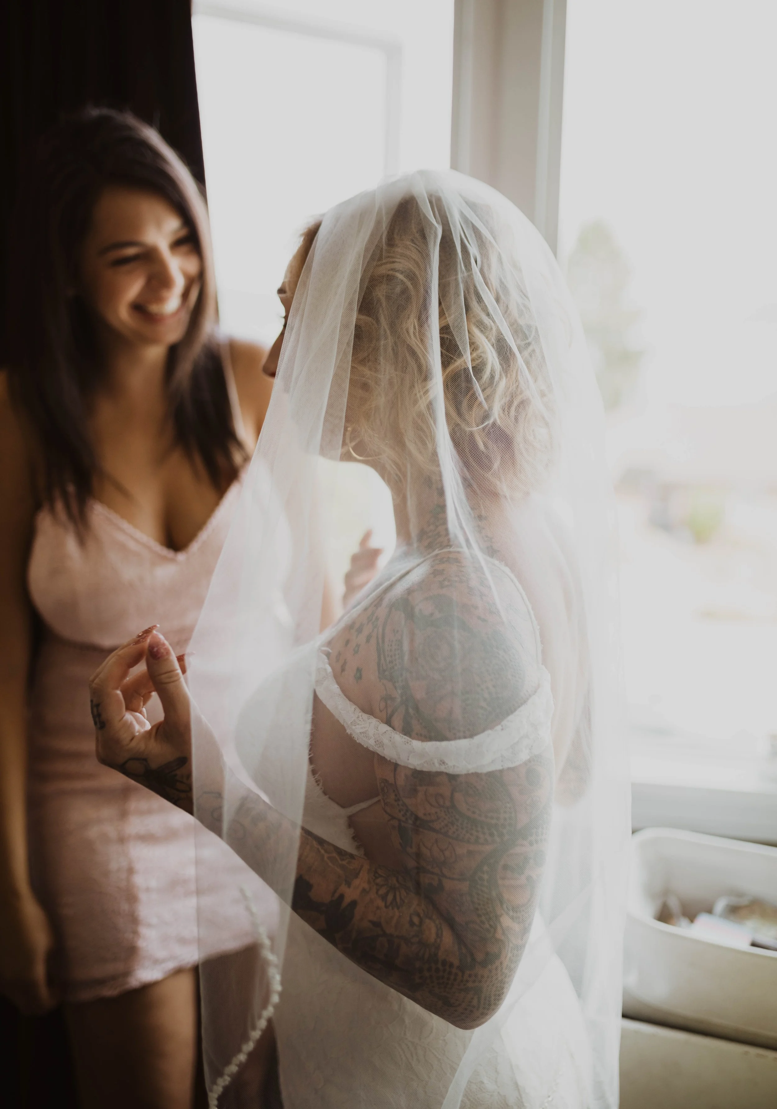 A bride with tattoos and blonde hair under a veil, smiling and holding the veil, while a woman in pink pajamas smiles in the background. Seattle, WA wedding photography.