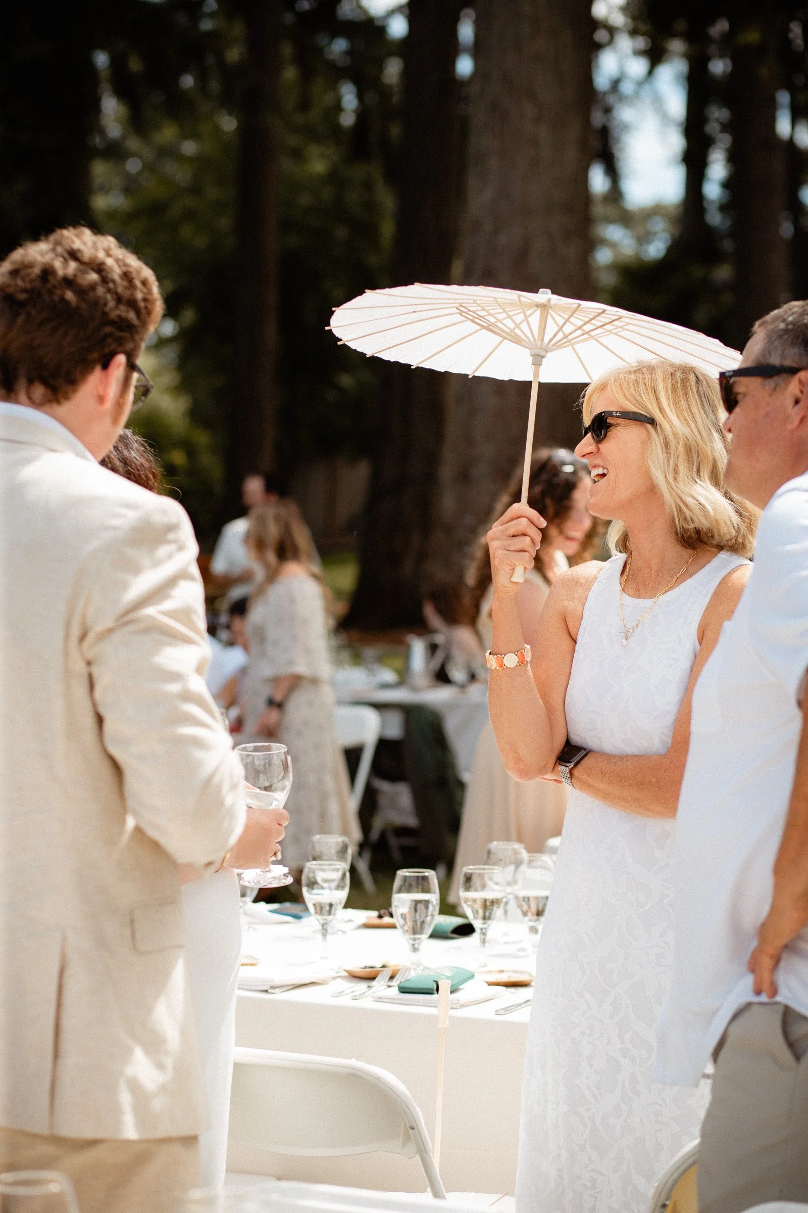 Wedding guests mingle at a table together holding umbrellas for shade at this lakeside wedding in Seattle, WA