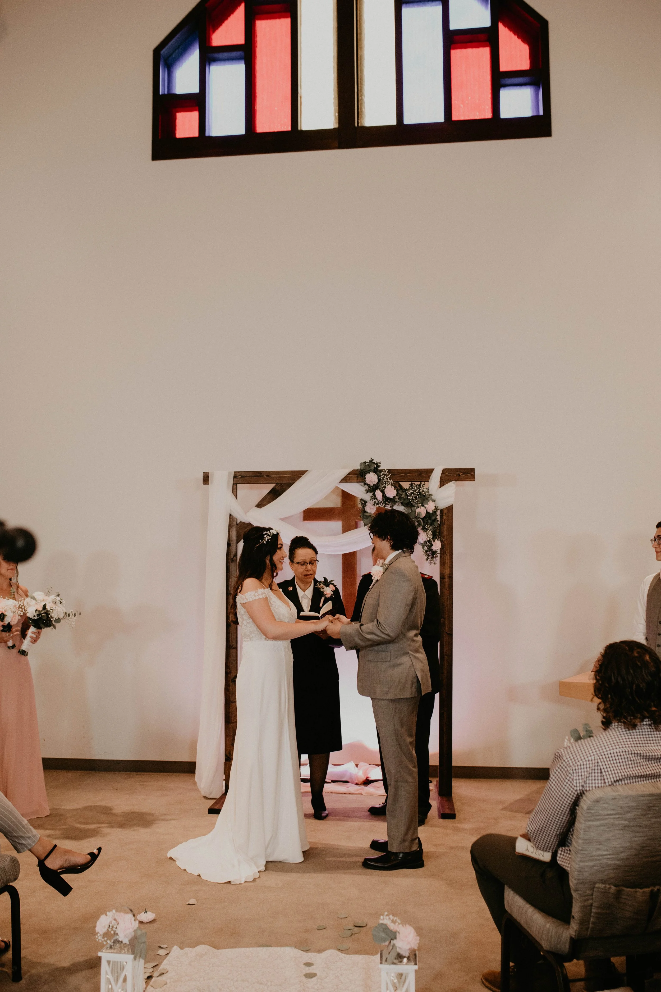 A couple getting married during a ceremony in a room with a stained glass window at the top. The bride is in a white gown, and the groom is in a gray suit. They are holding hands, facing each other, and exchanging vows or rings. An officiant stands b