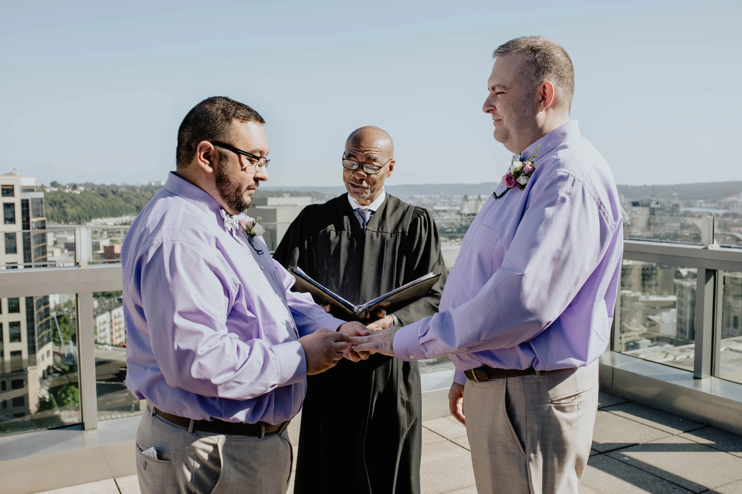 Two men in dress shirts and boutonnieres hold hands during a wedding ceremony on a rooftop with a cityscape in the background, officiated by a person in a judicial robe. Seattle Municipal Courthouse wedding photography.