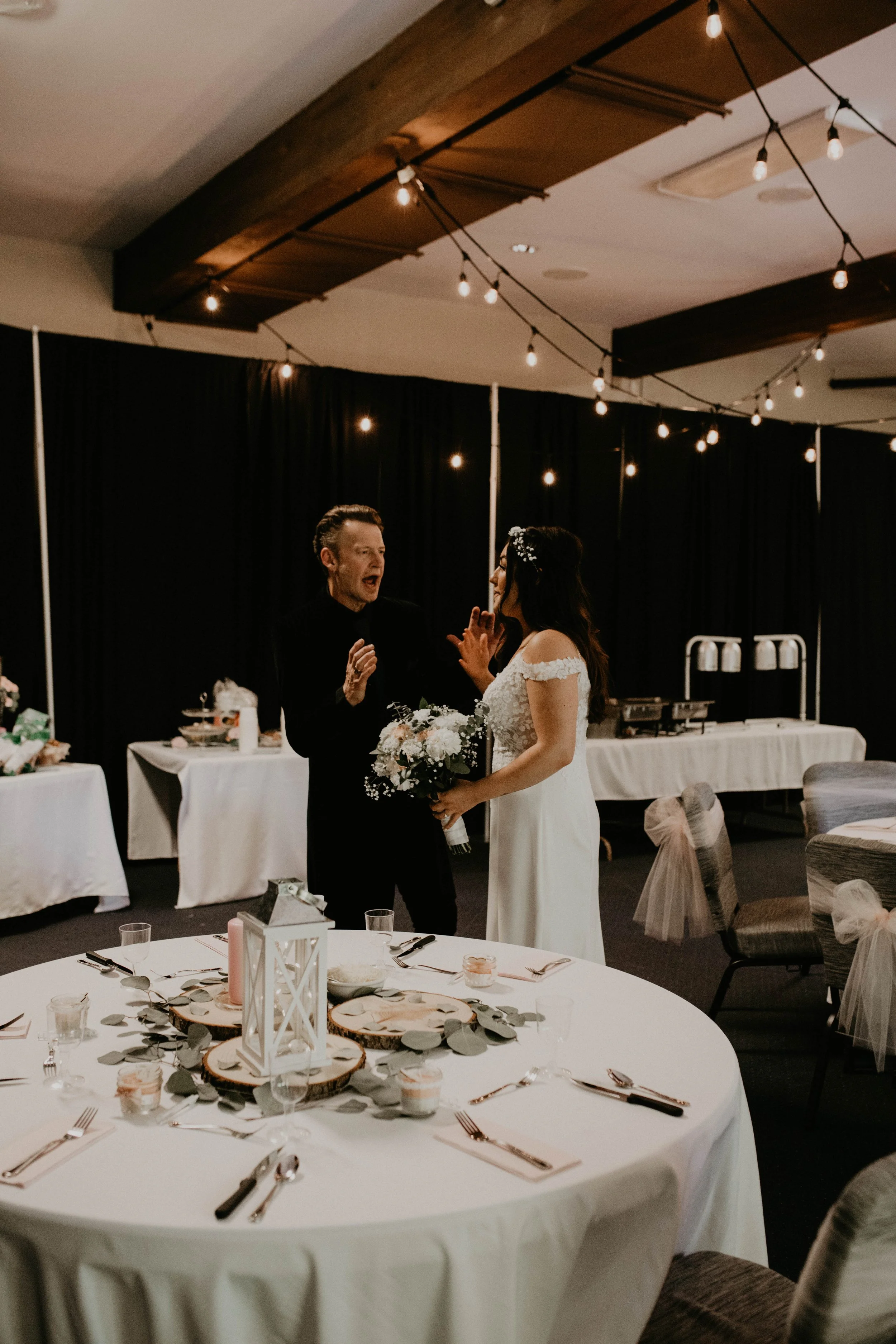 A bride in a white wedding dress holding a bouquet, talking to a man in a black suit at a wedding reception in a decorated banquet hall with string lights and round tables. Seattle, WA wedding photography.