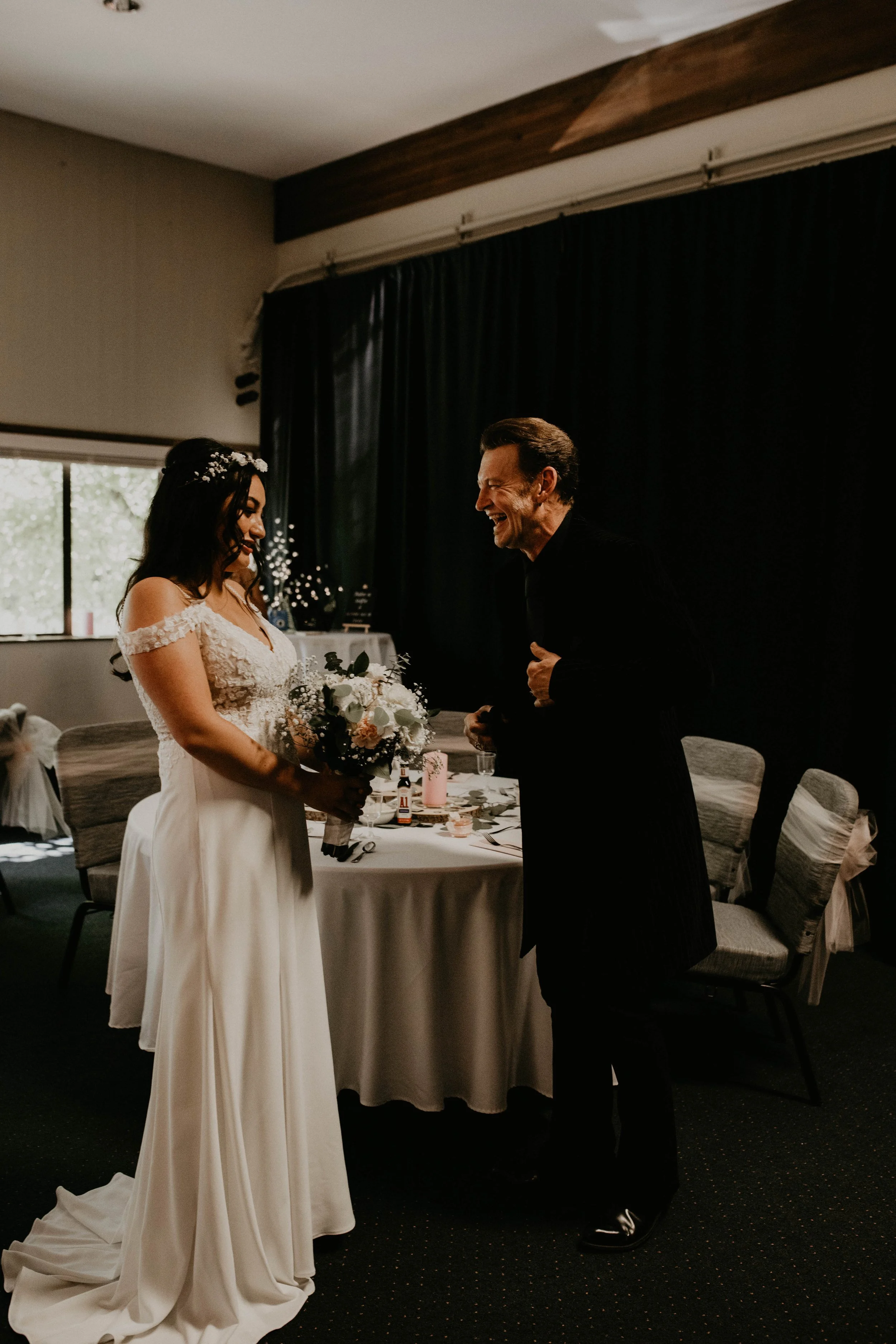 A bride holding a bouquet and a groom sharing a happy moment inside a room with dark curtains and chairs. Seattle, WA wedding photography.